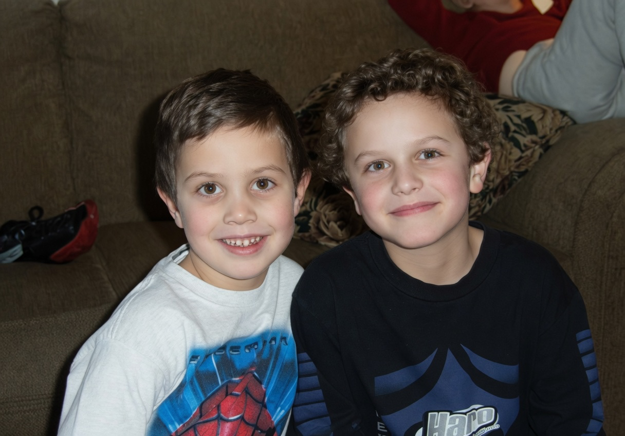 Two young boys sitting on a couch, smiling at the camera, one wearing a Spider-Man shirt and the other in a black shirt with a blue design.