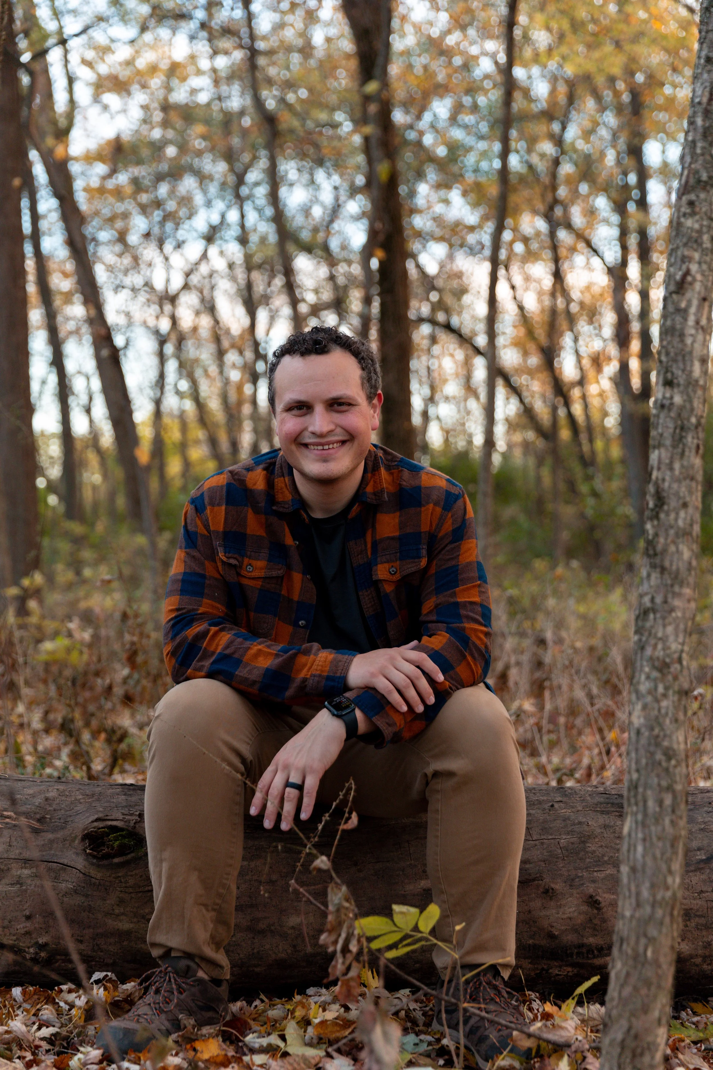 A man smiling, sitting on a fallen log in a forest during autumn with trees and fallen leaves in the background.