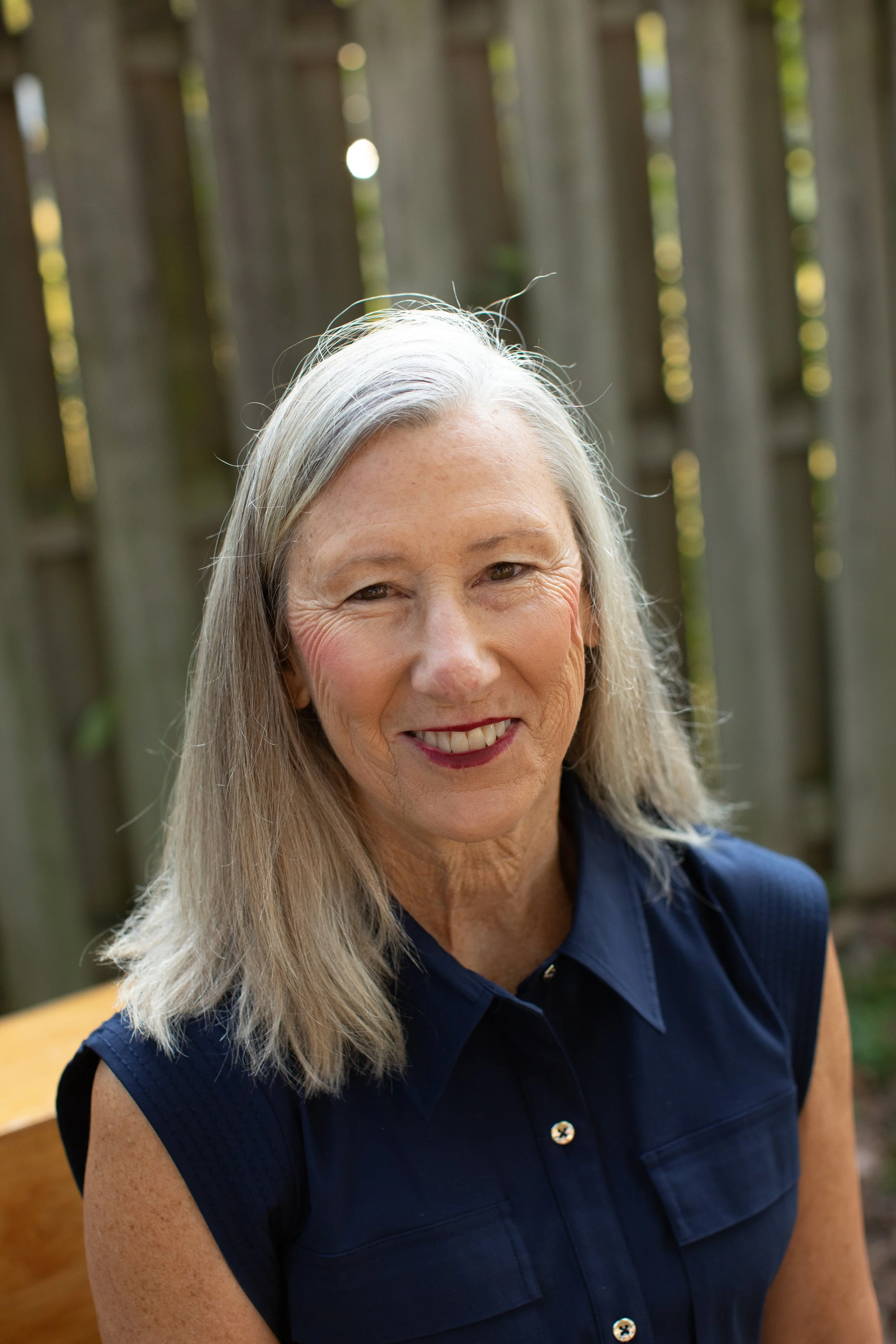 A smiling mature woman with long gray hair, wearing a sleeveless navy blue top, sitting outdoors in front of a wooden fence with bokeh sunlight.