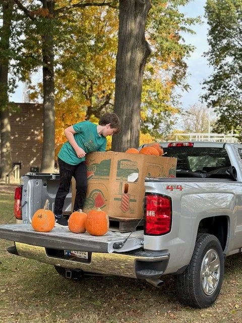 A boy loading pumpkins from a cardboard box into the back of a gray pickup truck parked outdoors during fall.
