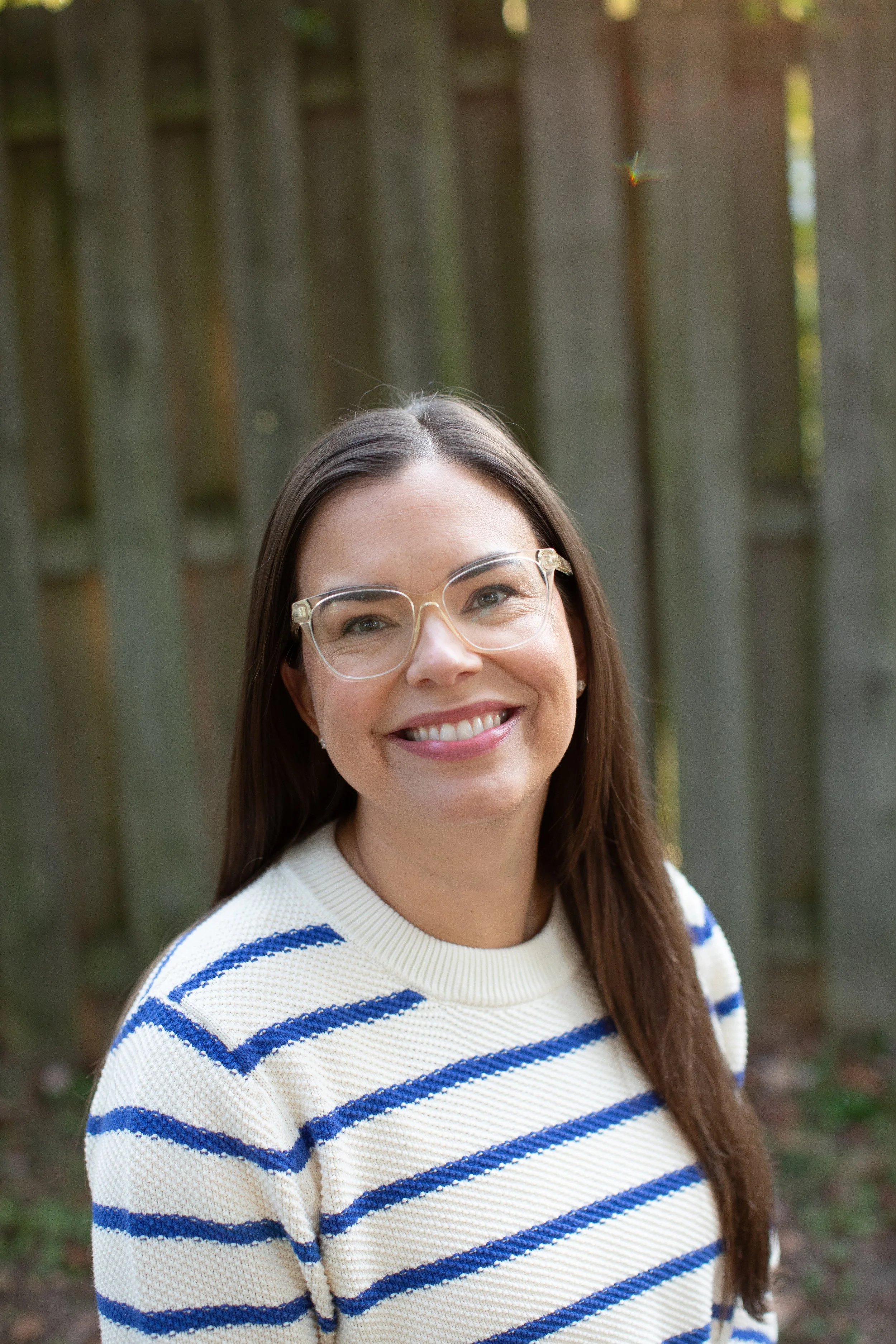 Smiling woman with long brown hair, wearing clear glasses and a white sweater with blue stripes, standing outdoors with a wooden fence in the background.
