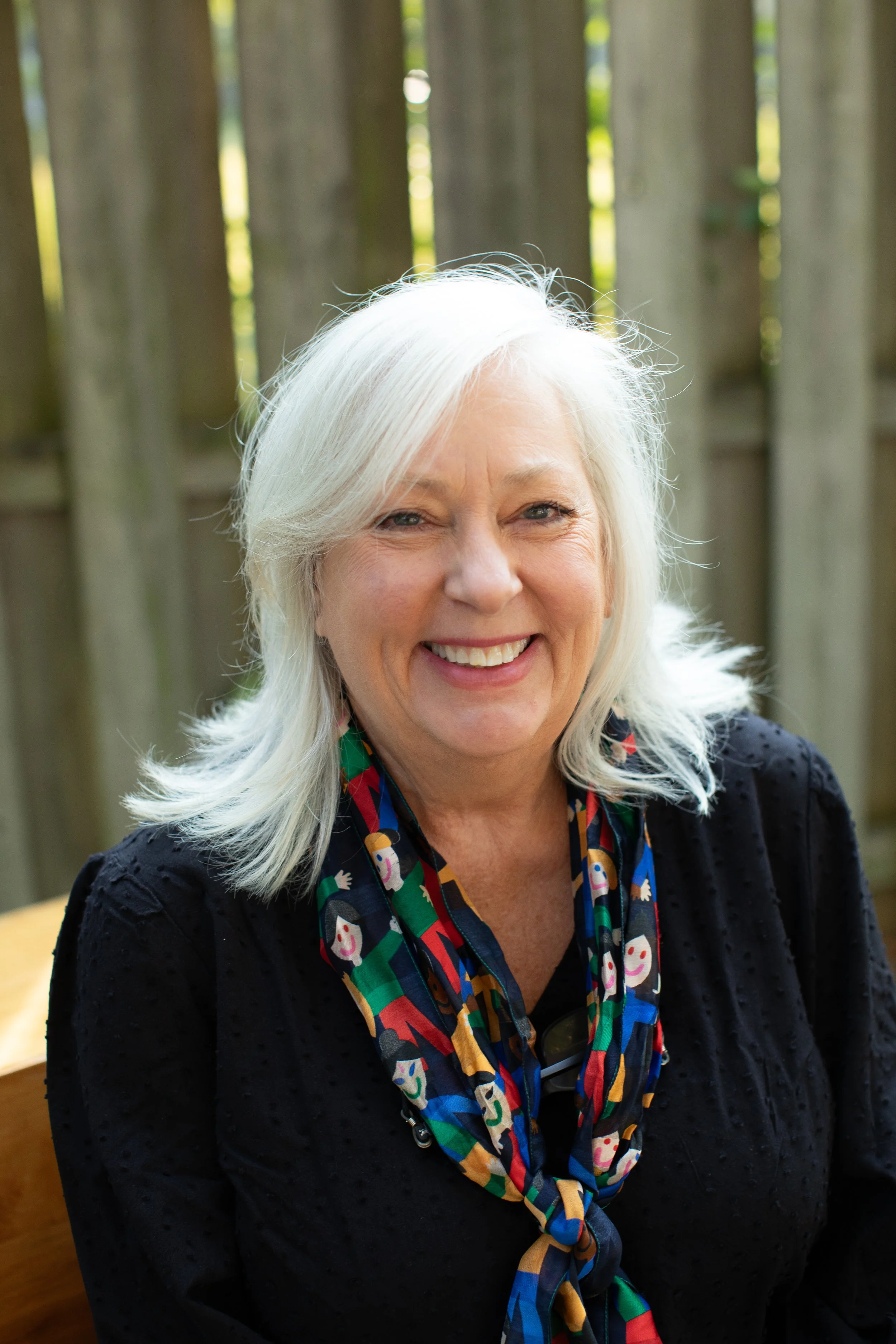 A smiling woman with long white hair wearing a colorful scarf and black top, sitting outdoors near a wooden fence.