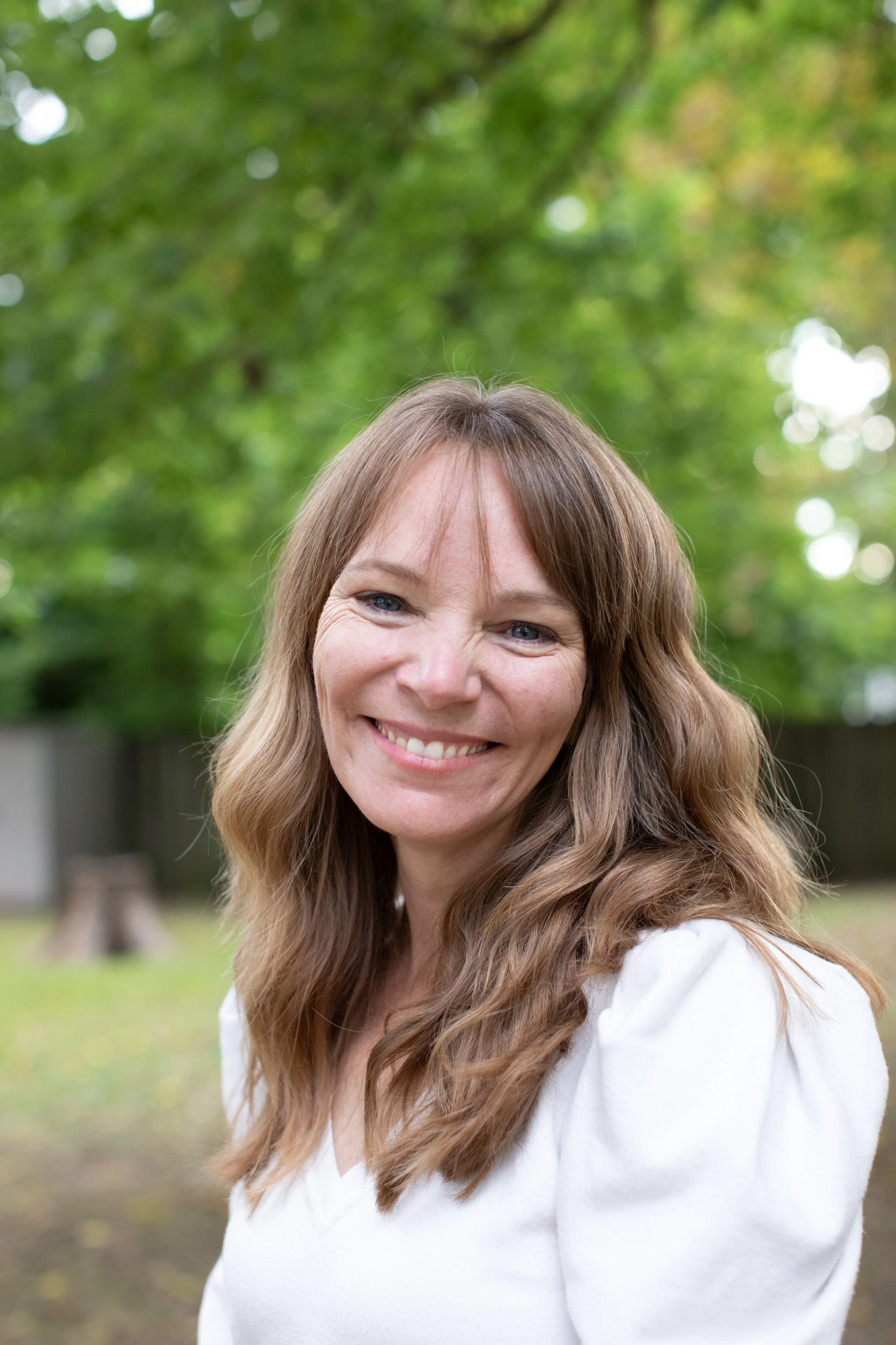 Smiling woman with long wavy brown hair wearing a white top outdoors in a green park with trees in the background.