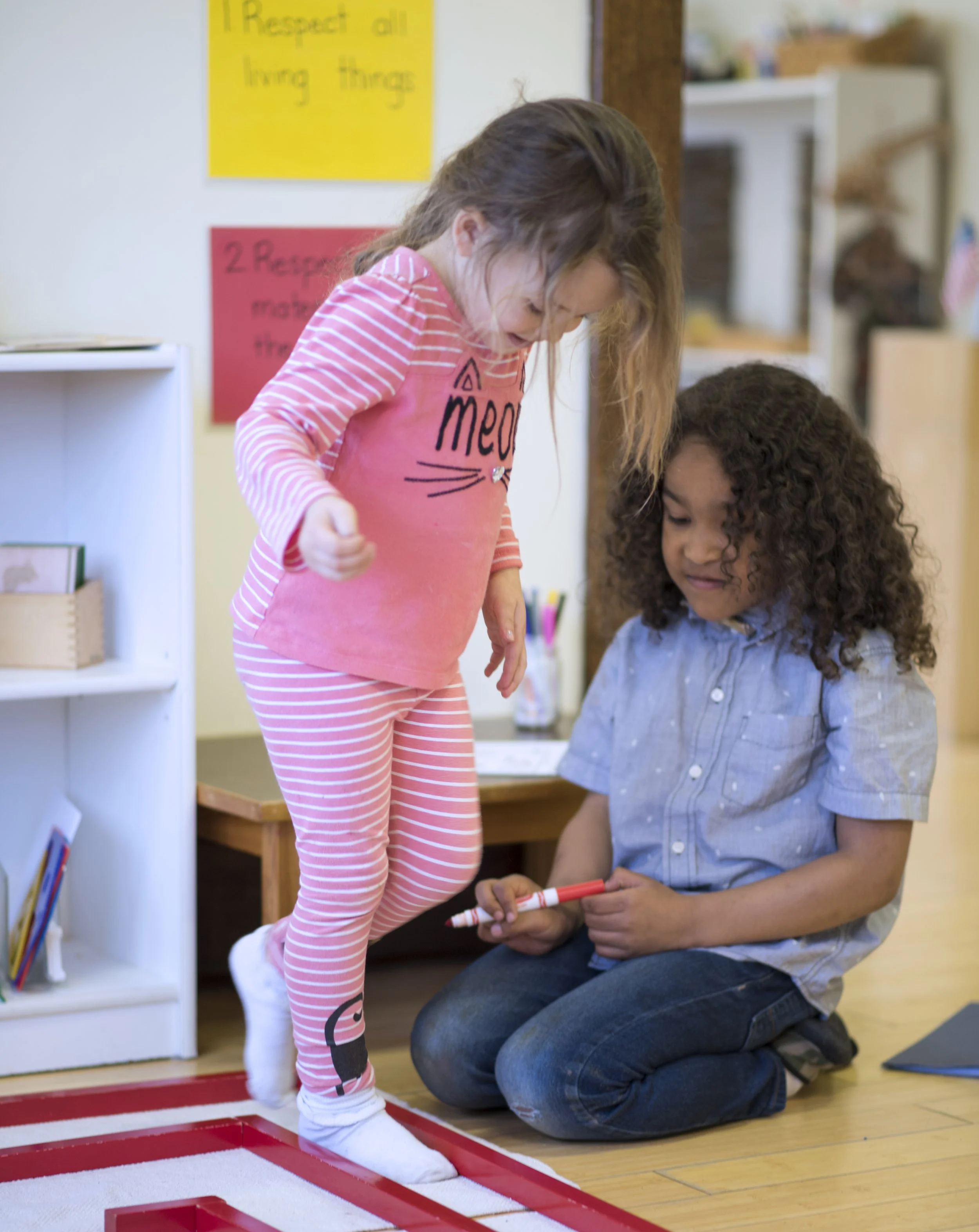 A young girl standing on a red and white taped boundary line while an adult man kneeling nearby helps her with a task using a red marker. The girl is wearing pink and white striped pajamas and white socks, and the man is dressed in a light blue shirt and dark jeans. They are in a room with bookshelves, a yellow and red poster on the wall, and a hardwood floor.