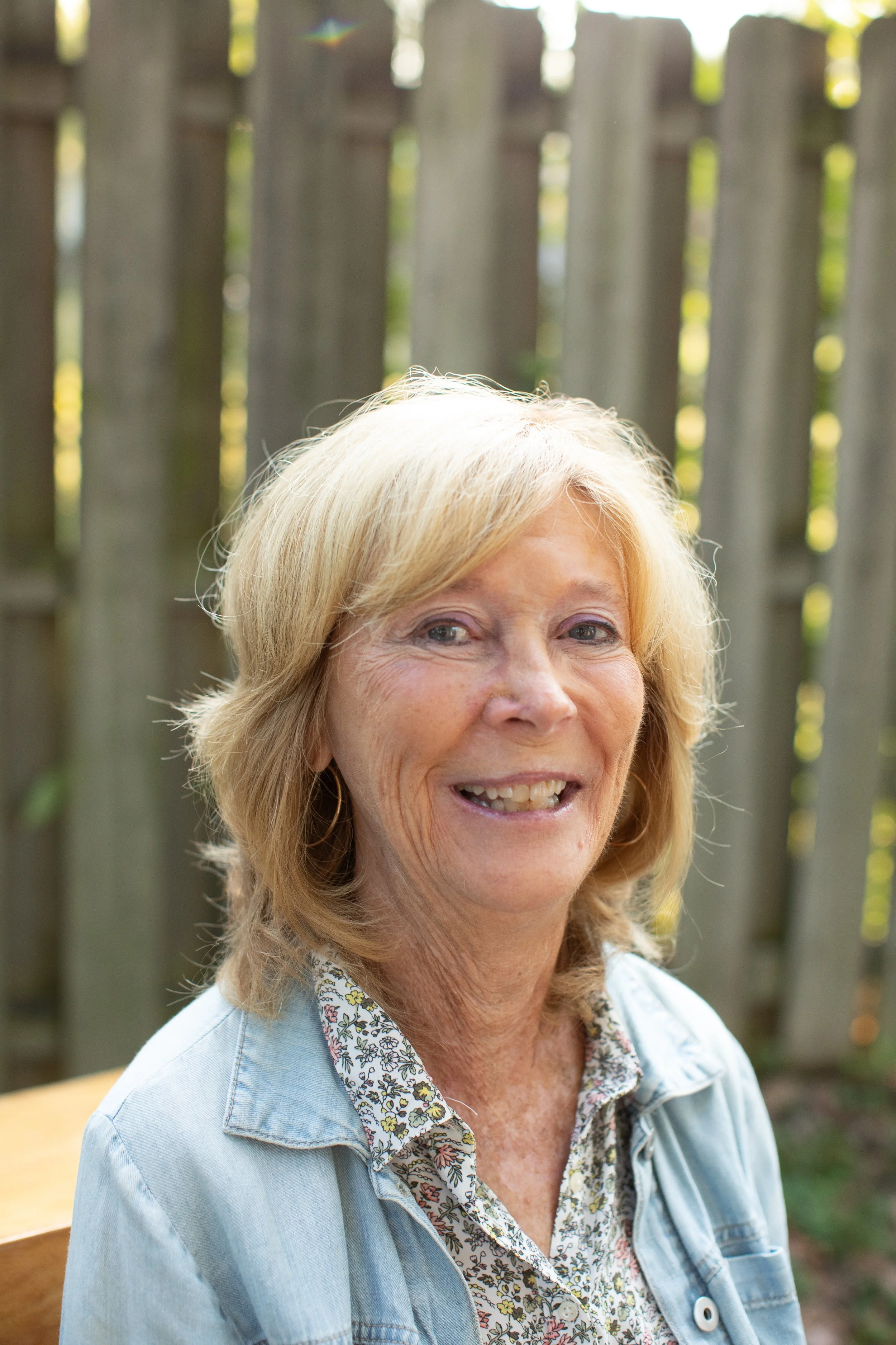 A smiling elderly woman with blonde hair, wearing a floral shirt and denim jacket, sitting outdoors in front of a wooden fence.