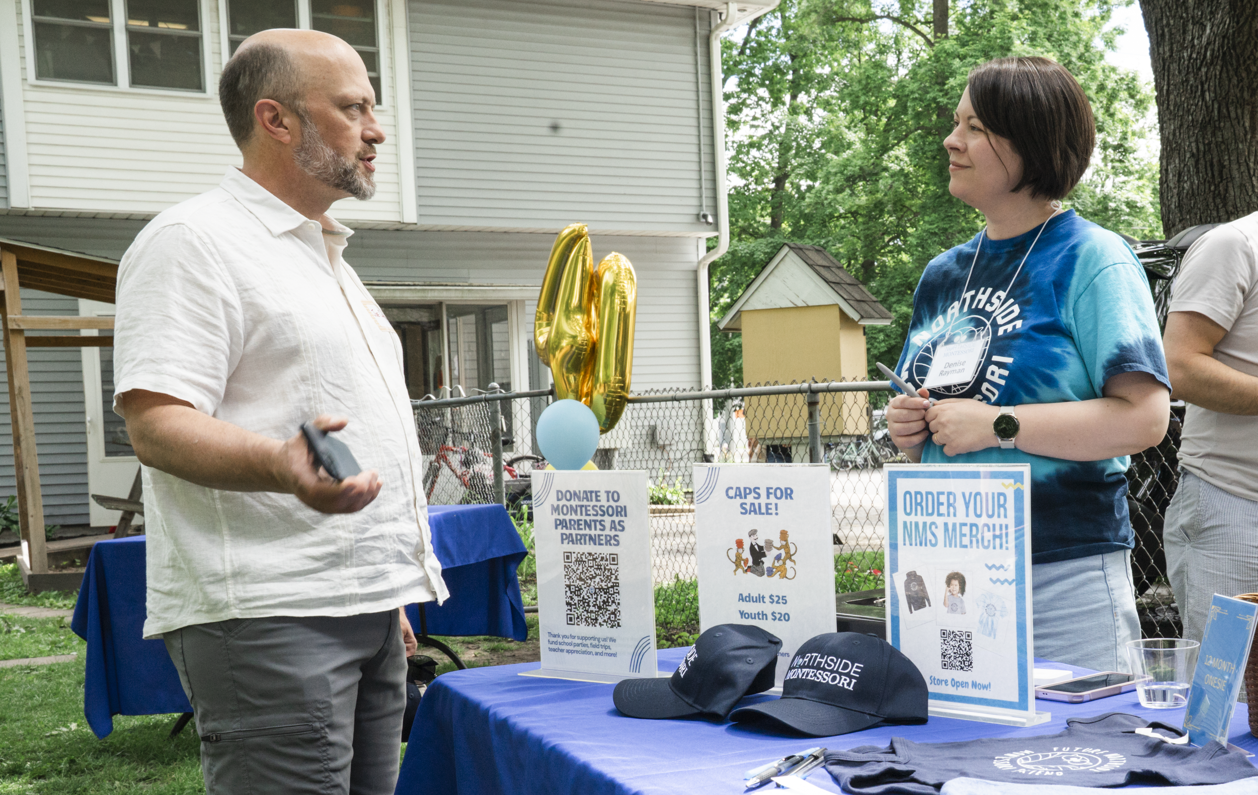 A man and a woman are having a conversation at an outdoor event booth. The booth has signs with information about donations and merchandise, including a blue tablecloth, hats, and promotional materials. The woman is wearing a blue T-shirt with a logo and a name tag, and the man is holding a phone. There are green trees and a house in the background.