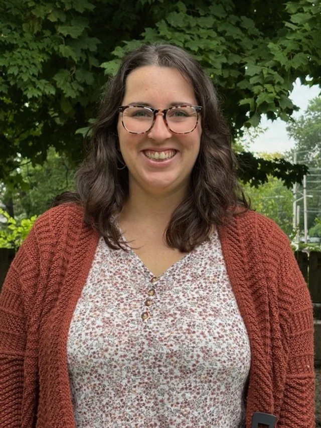 A young woman with shoulder-length curly brown hair, glasses, and a big smile, standing outdoors in front of green trees, wearing a brownish-red knitted cardigan over a floral blouse.