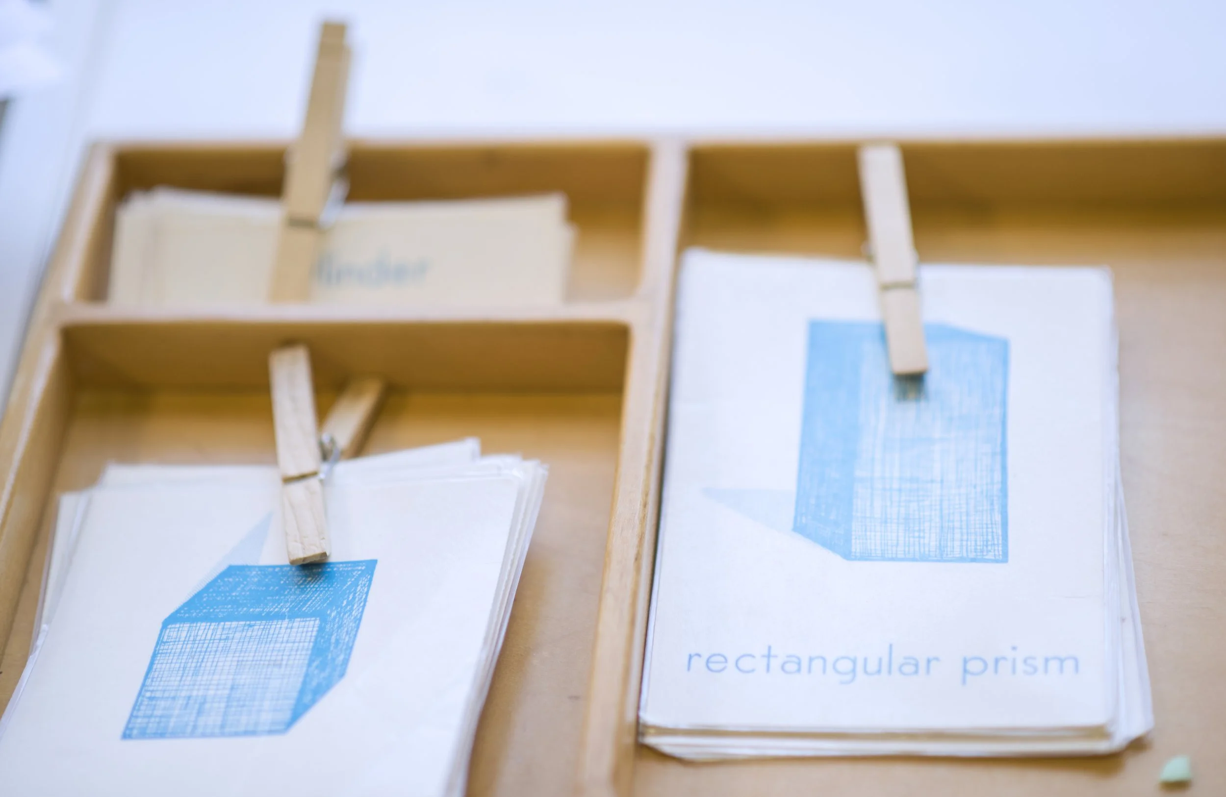 Close-up of educational materials with blue illustrations of rectangular prisms and some paper clips, on a wooden surface.