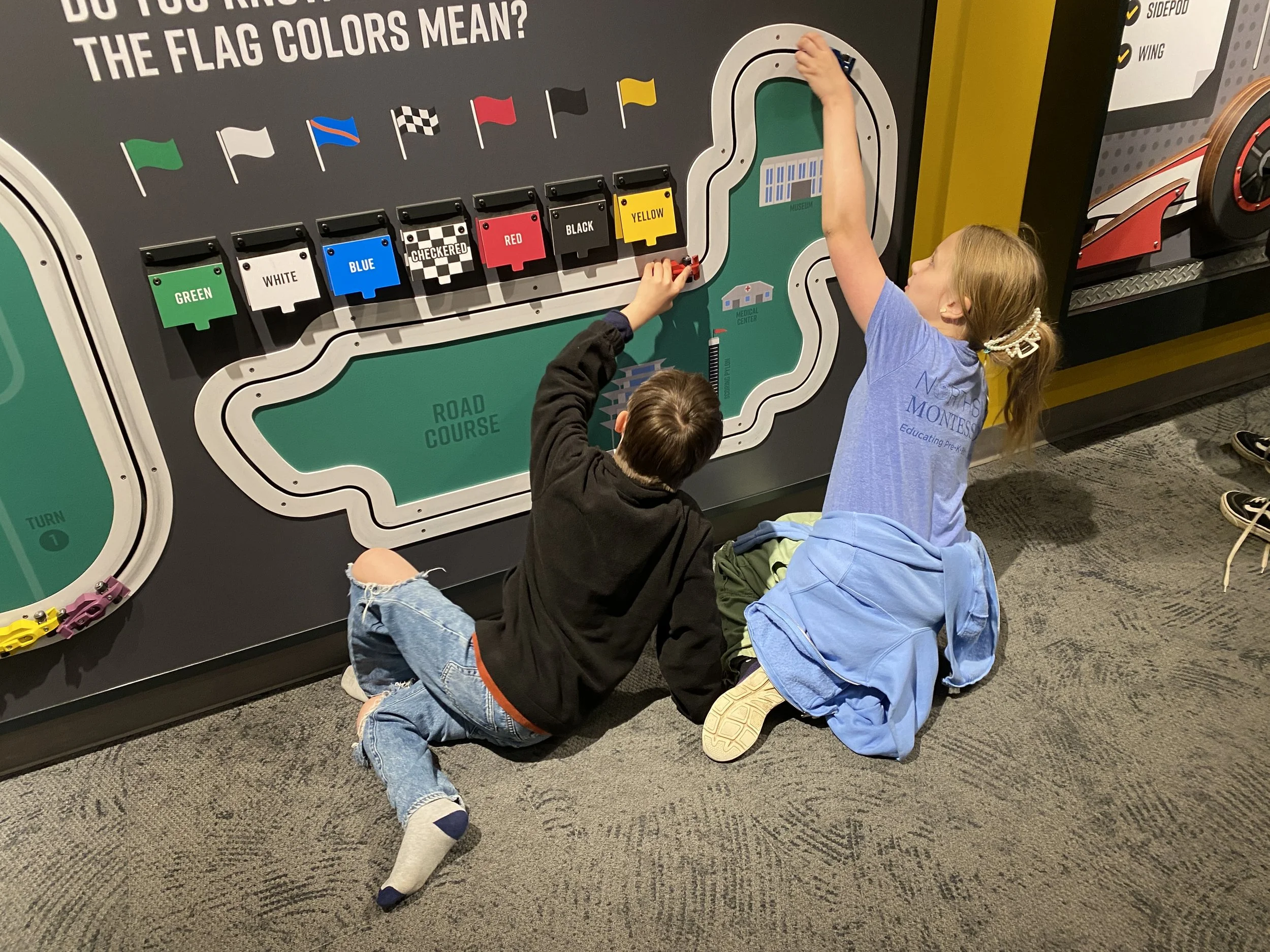 Three children sitting on dark carpeted floor playing with a large wall-mounted train control panel at a museum or interactive exhibit. The control panel has flags, buttons, and display labels related to a train theme.