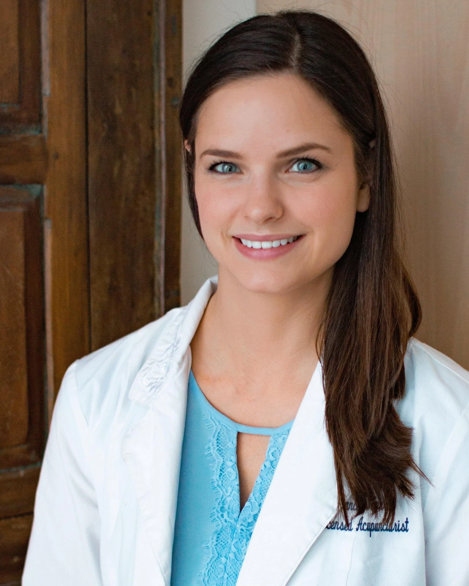 A woman with blue eyes and long brown hair wearing a white coat and blue scrubs standing in front of a wooden wall, smiling at the camera.