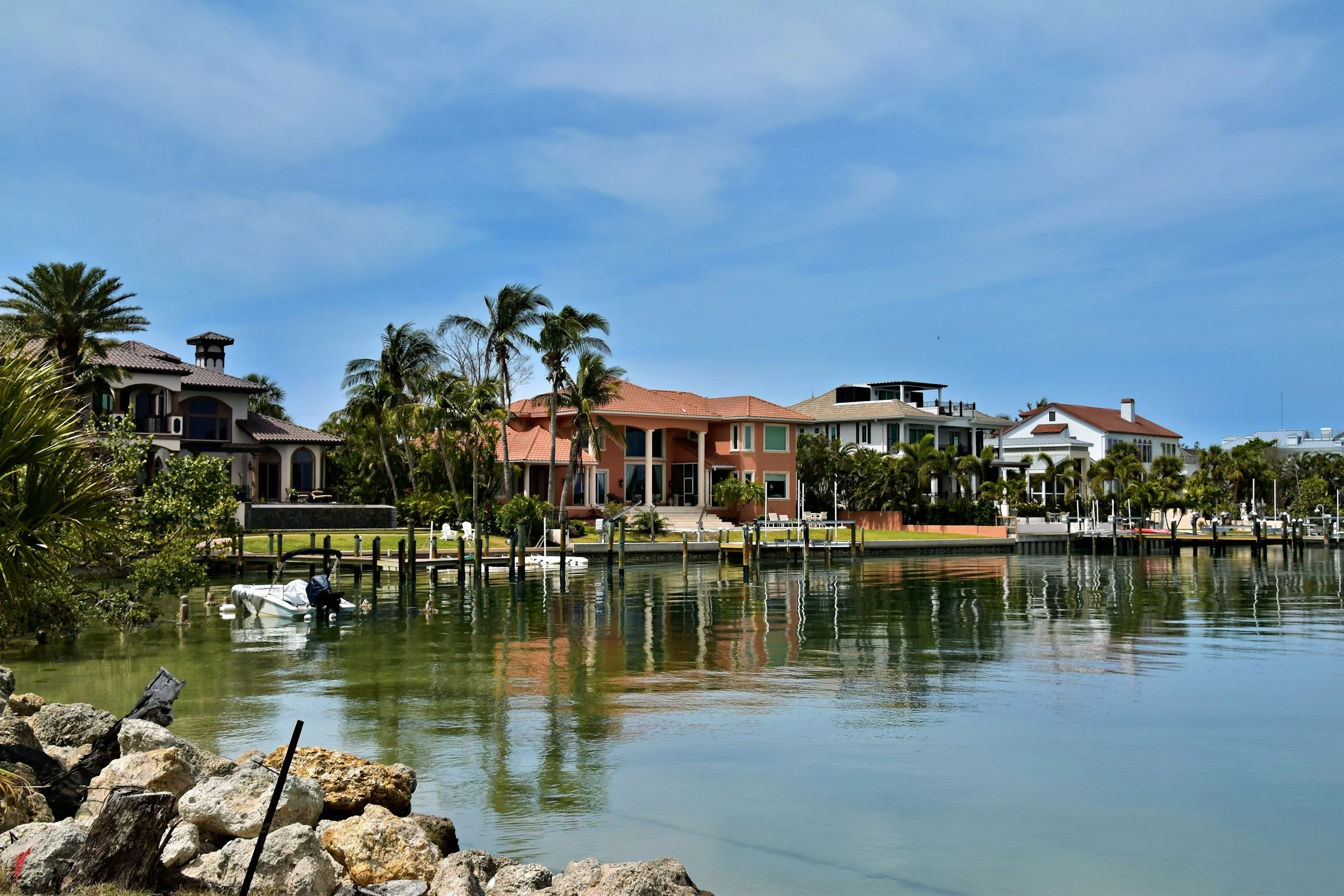 Waterfront houses with palm trees along the water, boats docked at the piers, rocks in the foreground, and a partly cloudy sky.