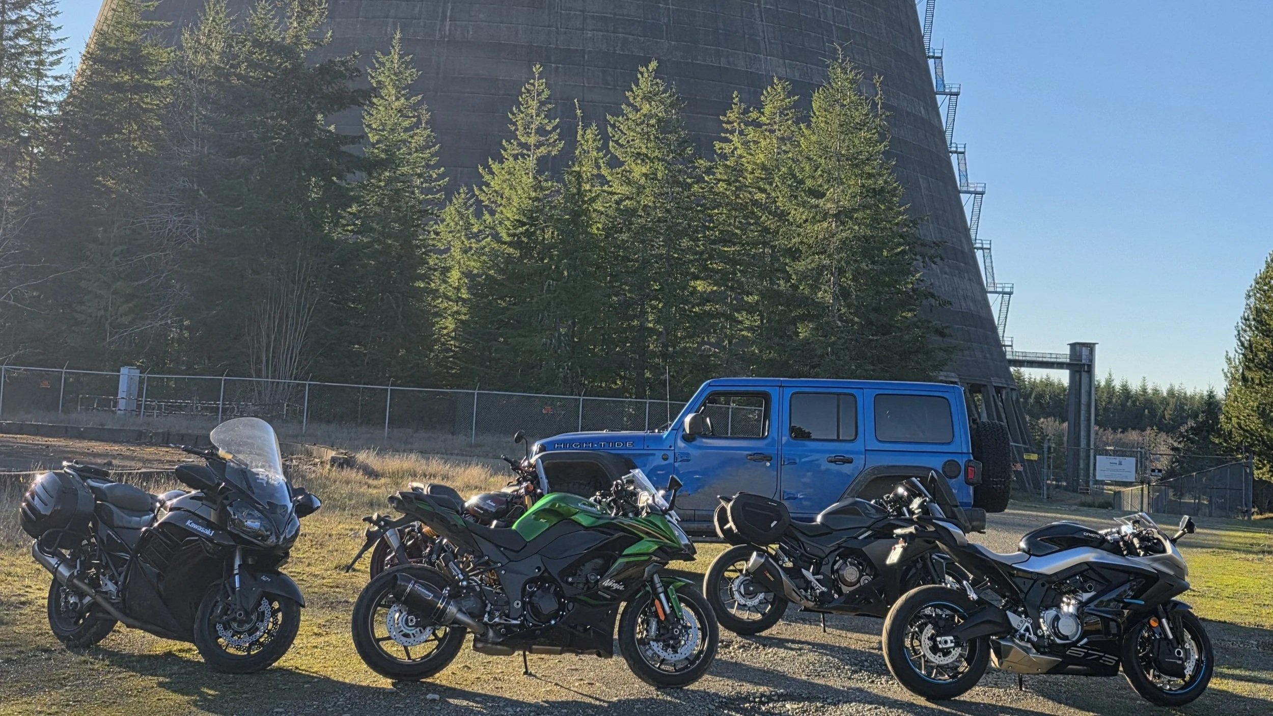 Several motorcycles parked in a lot with a blue off-road SUV and a large cooling tower during a DFD CYCLES group ride from Repair shop in Kitsap County Washinton.