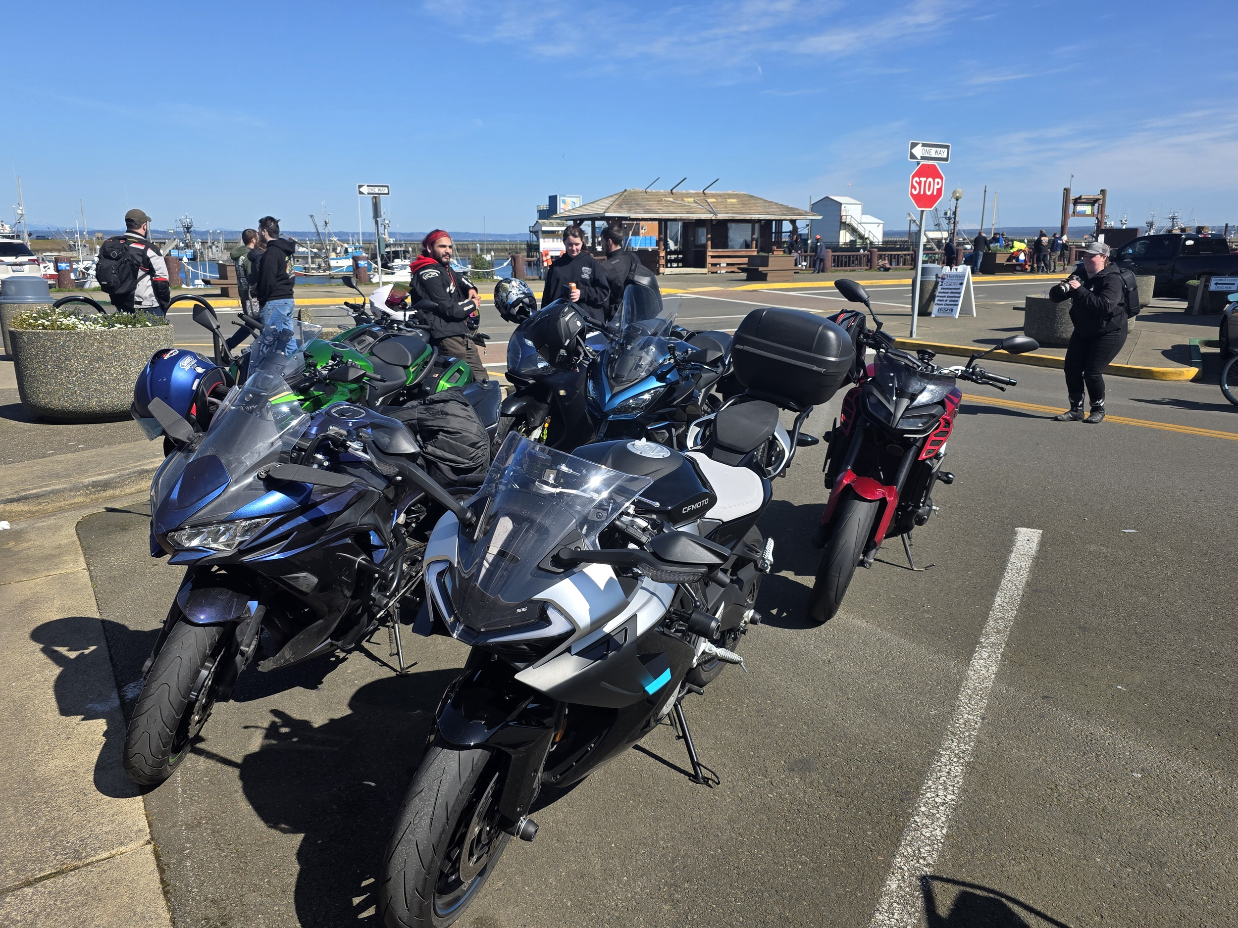 Multiple motorcycles parked in a lot near a waterfront, with people standing and talking, and a small building and boats visible in the background under a partly cloudy sky.