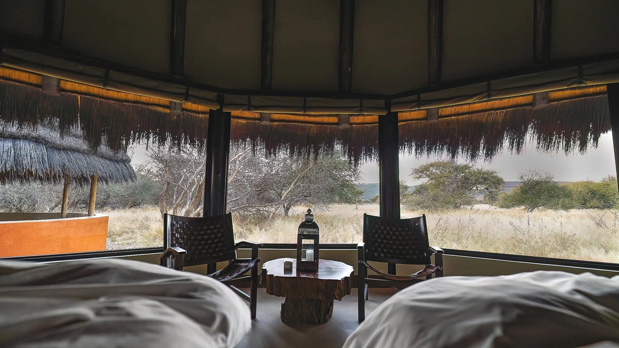 View from inside a room with large windows, looking out onto a savannah landscape with trees and dry grass. There are two chairs and a table with a lantern on it in the room.