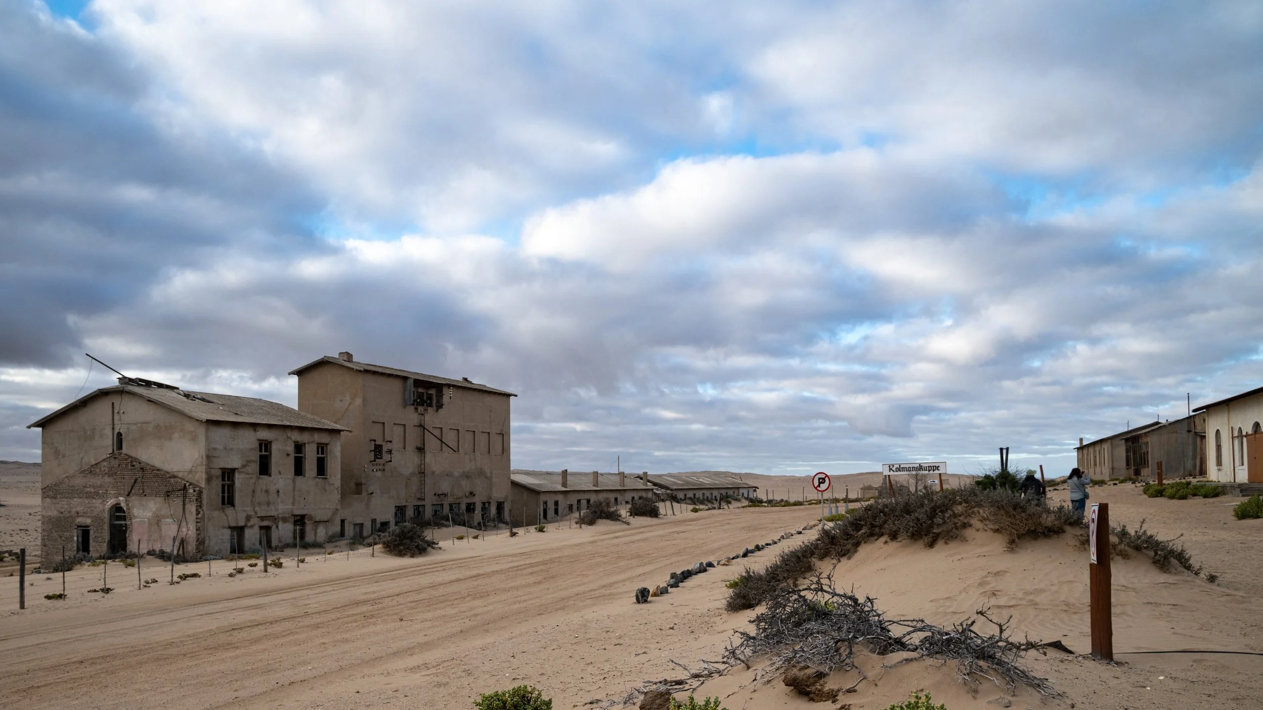 Old abandoned buildings in a desert landscape under a cloudy sky, with a sign that says 'Kolmanskuppe' and a few people walking nearby.