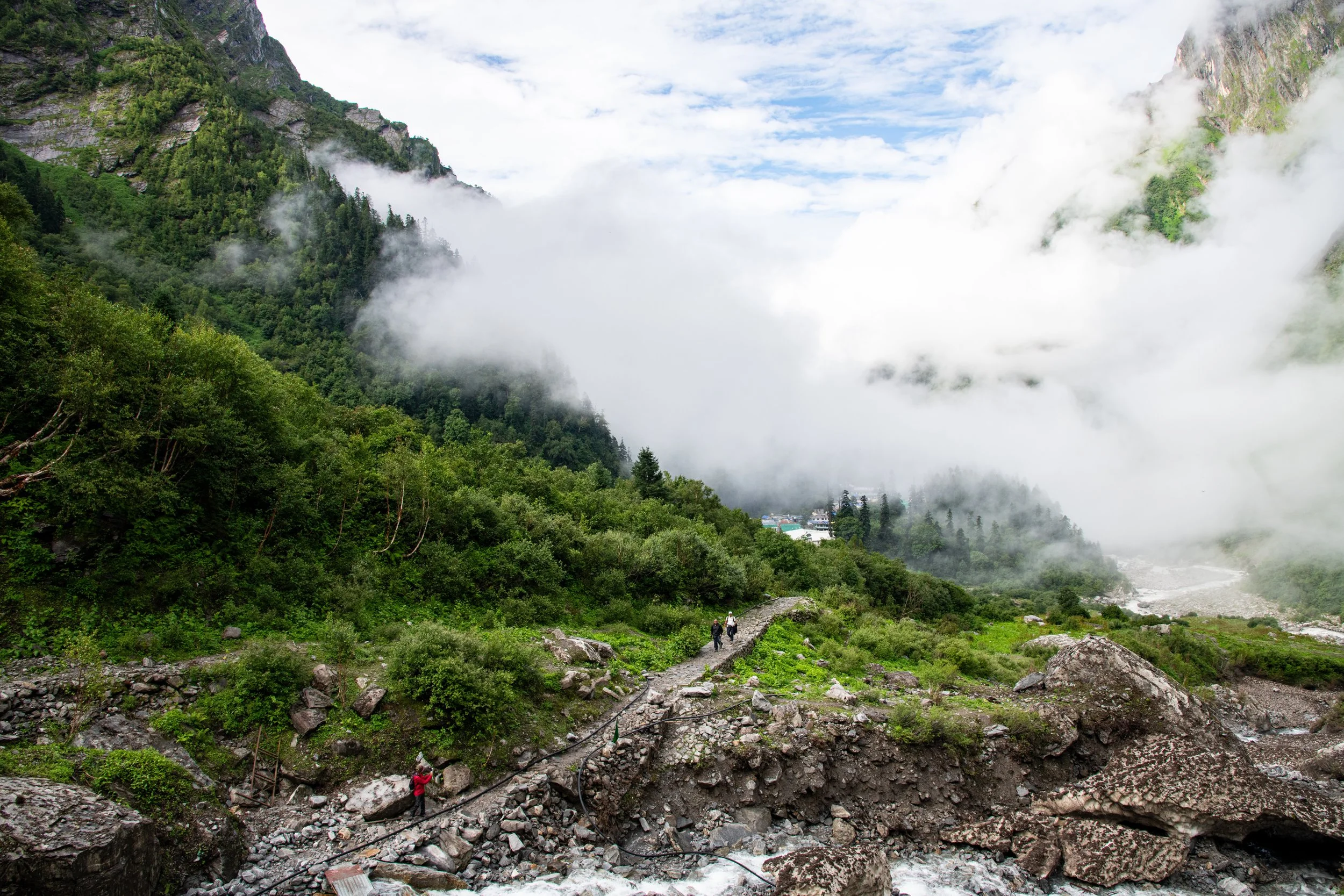 Two hikers walking along a rocky mountain trail with lush green trees on either side, misty mountains and clouds in the background.