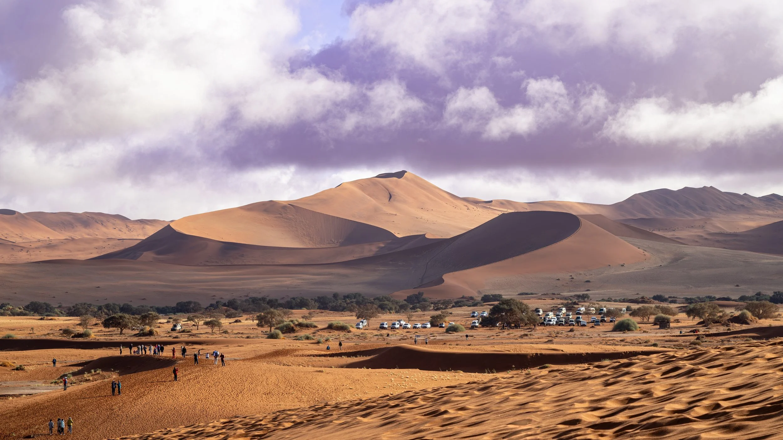 Sand dunes in a desert landscape with a mountain in the background and people walking in the foreground.