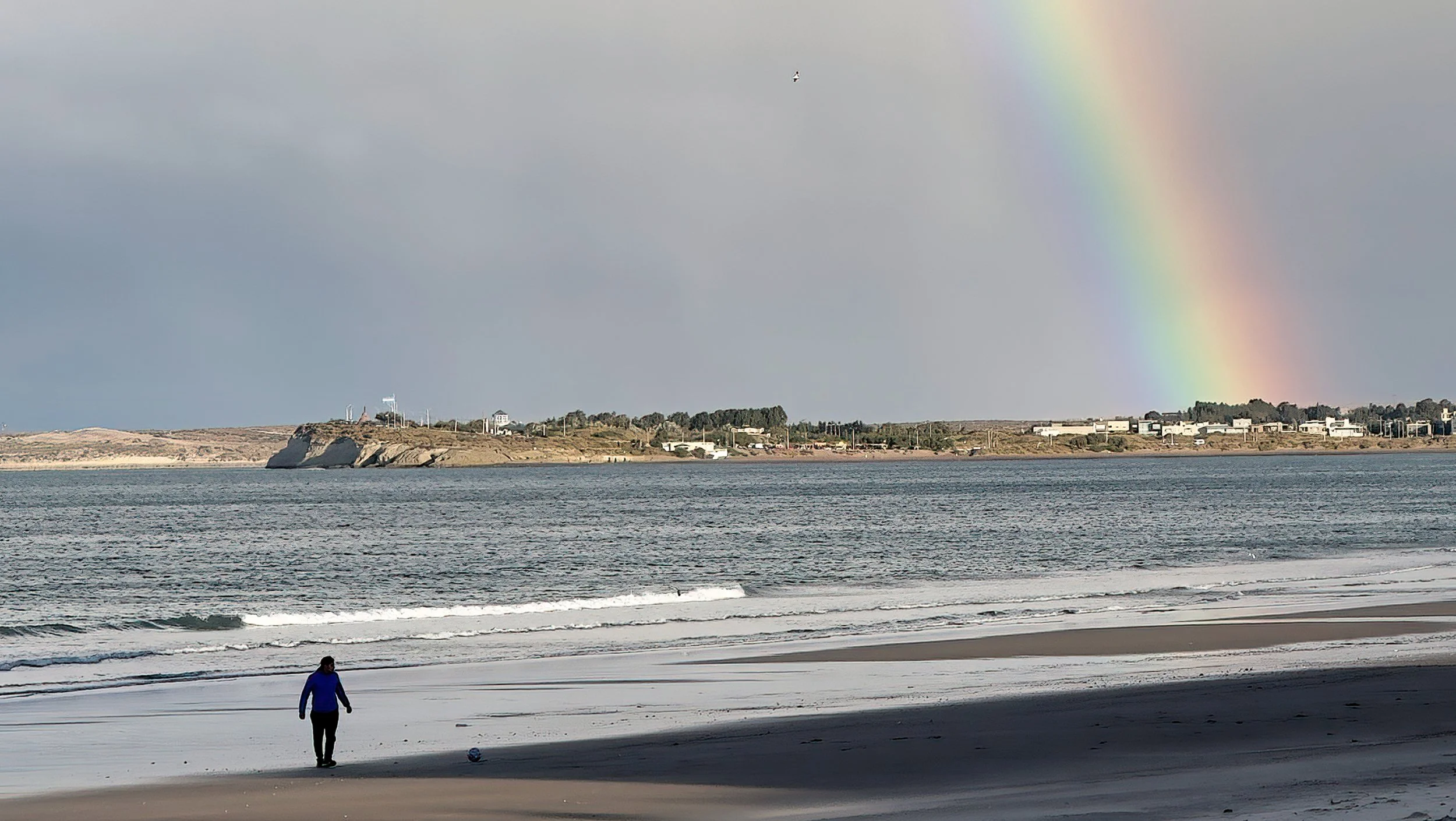 Person walking along the beach with a ball, ocean waves, distant shoreline with buildings, clouds, rainbow in the sky.