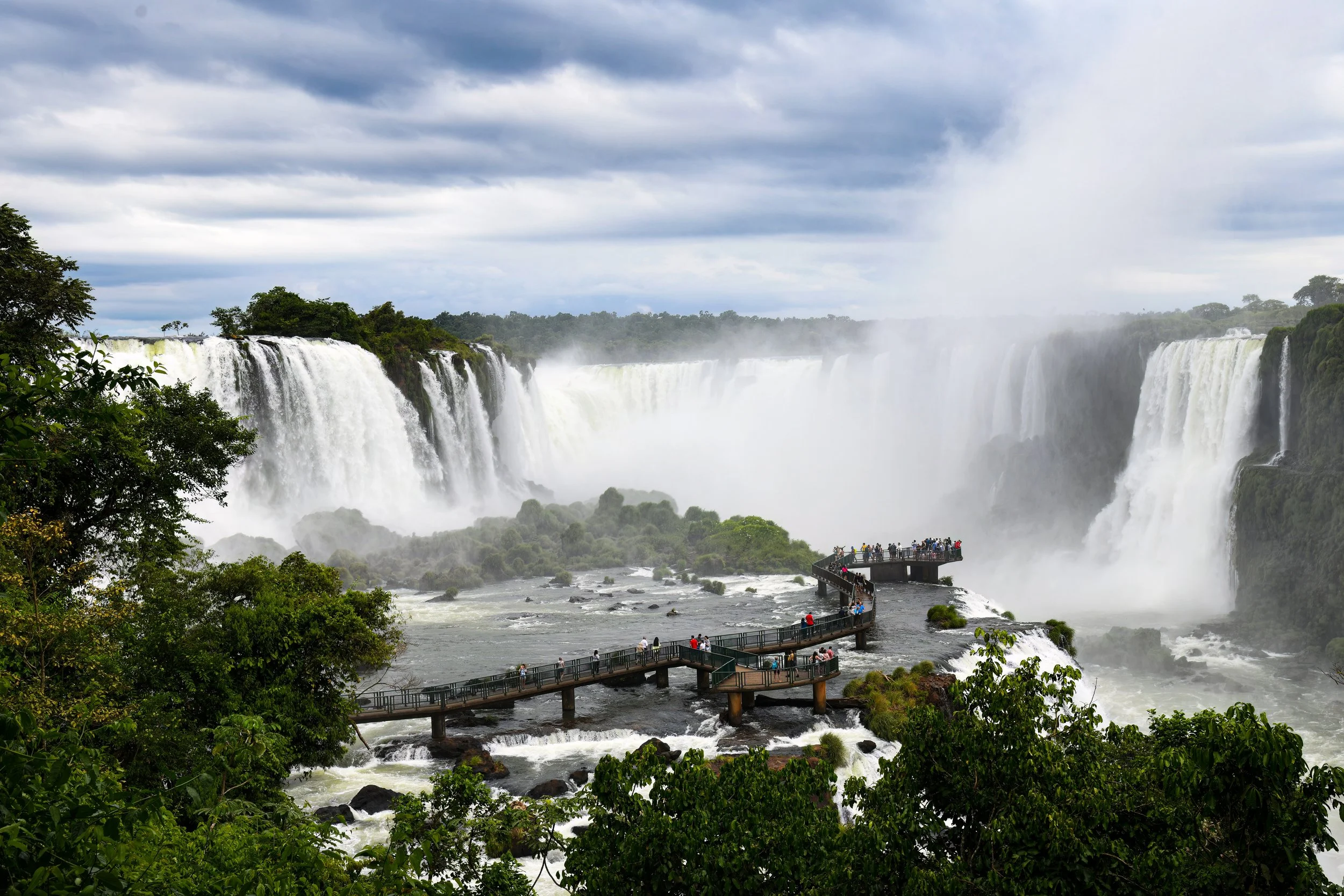 View of Iguazu Falls with a viewing platform and many visitors, trees, and cloudy sky in the background.
