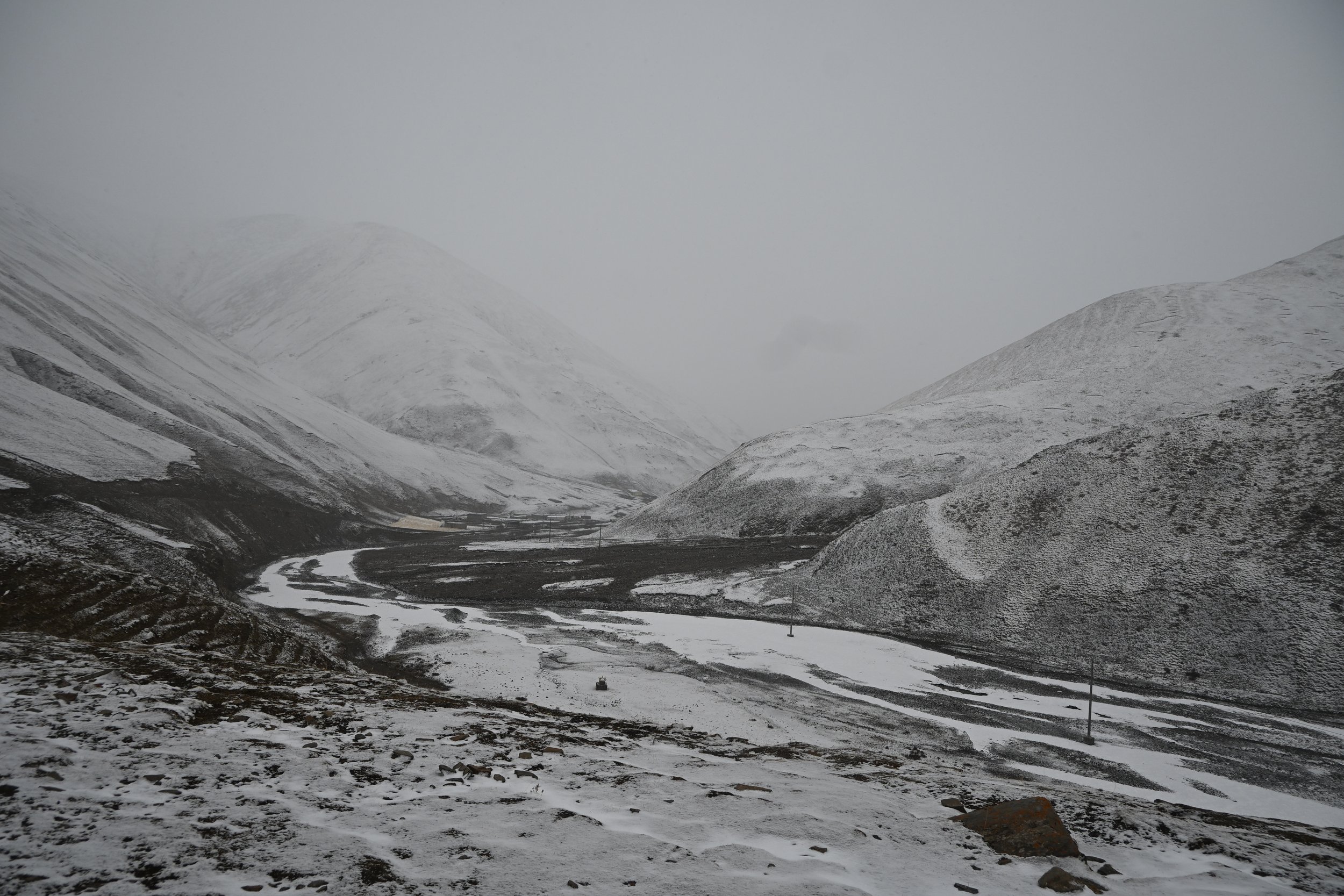 Snow-covered mountains with a winding partially frozen river in the valley below under a gray overcast sky.