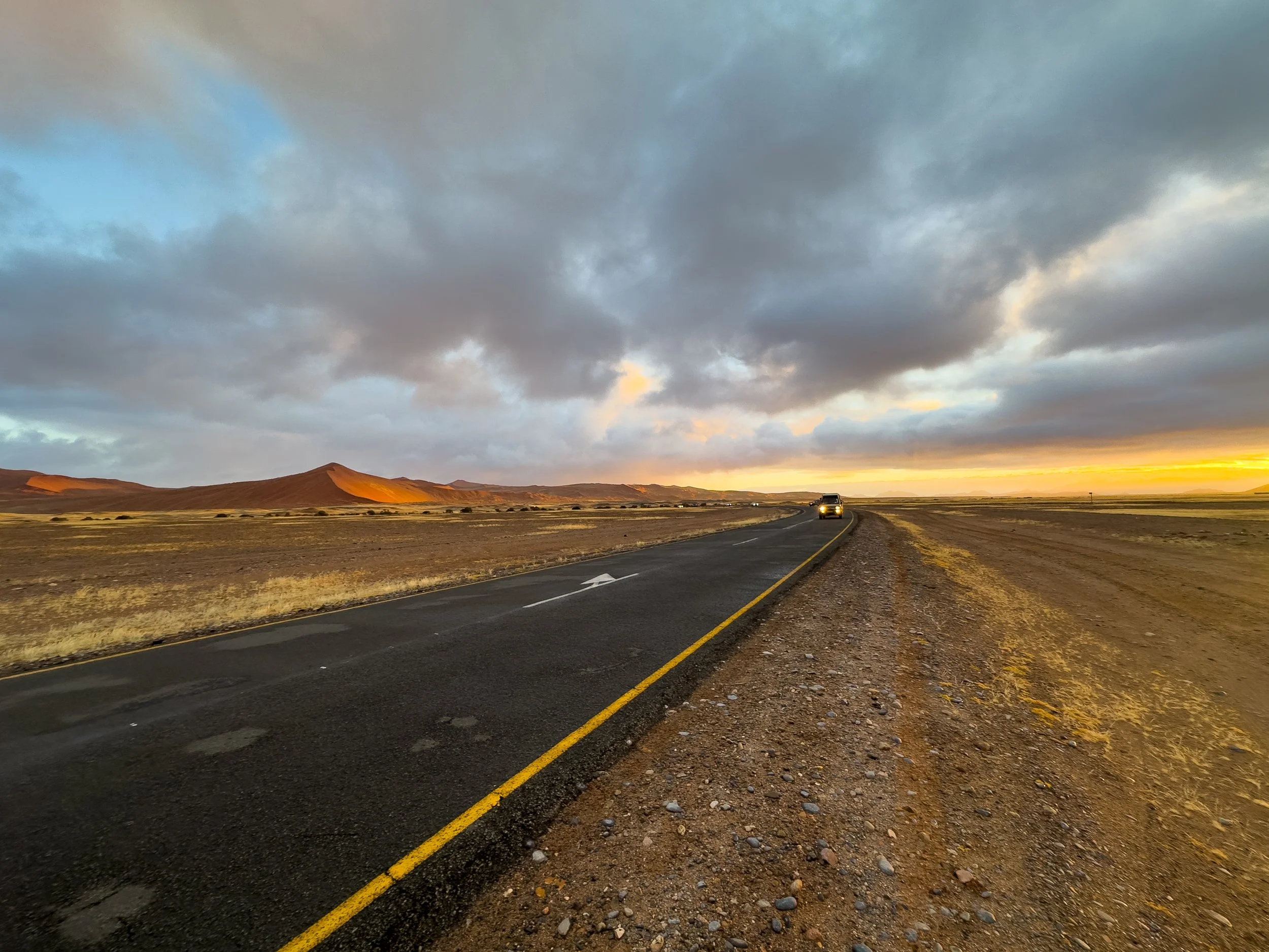 A car driving on a curved road through a desert landscape with rolling hills under a cloudy sky during sunset.