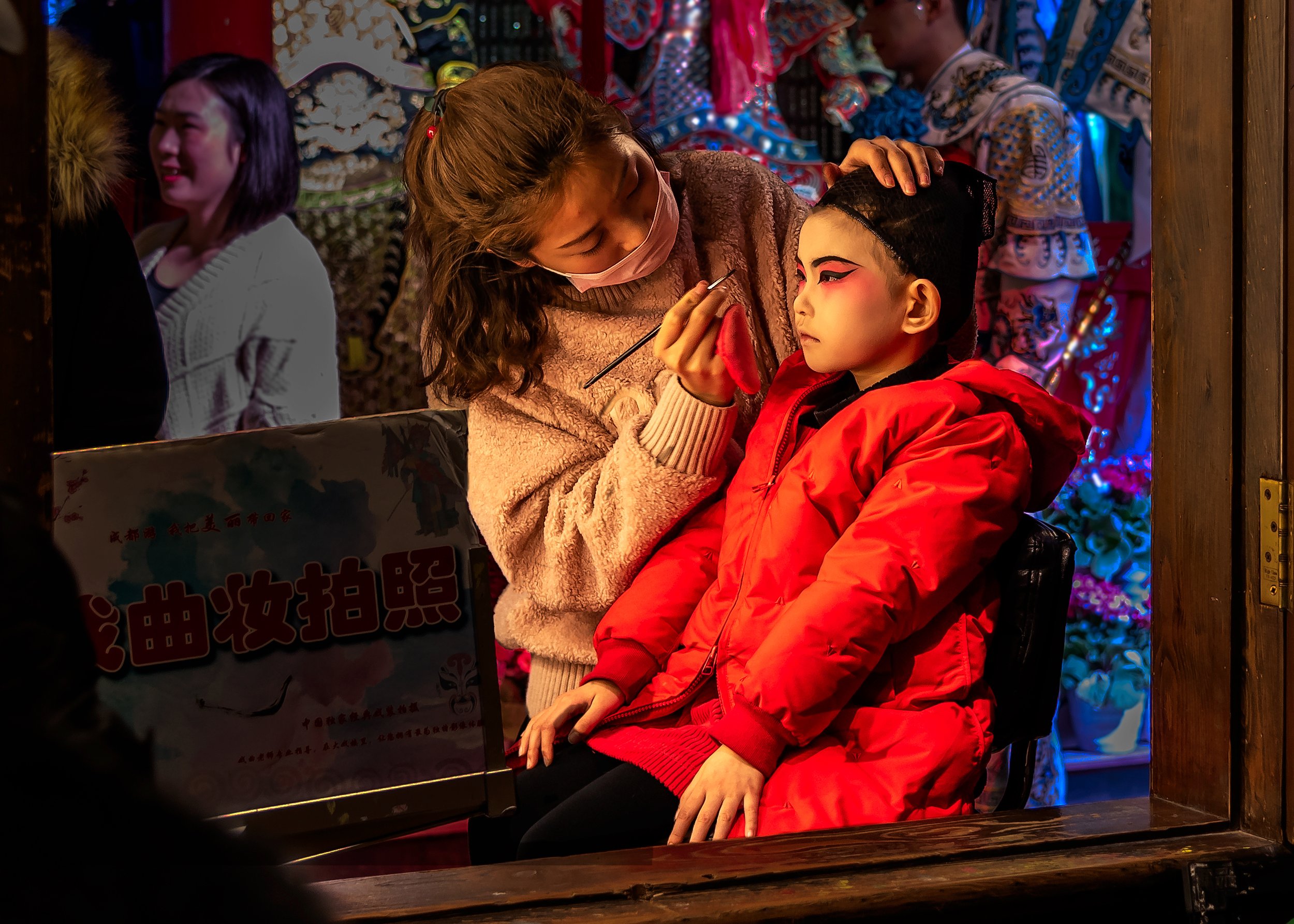 A young boy in a red jacket is getting his face painted by a woman with long brown hair, who is wearing a pink face mask and a beige sweater. They are sitting at a table with a colorful display in the background, and a young girl in white is smiling nearby.