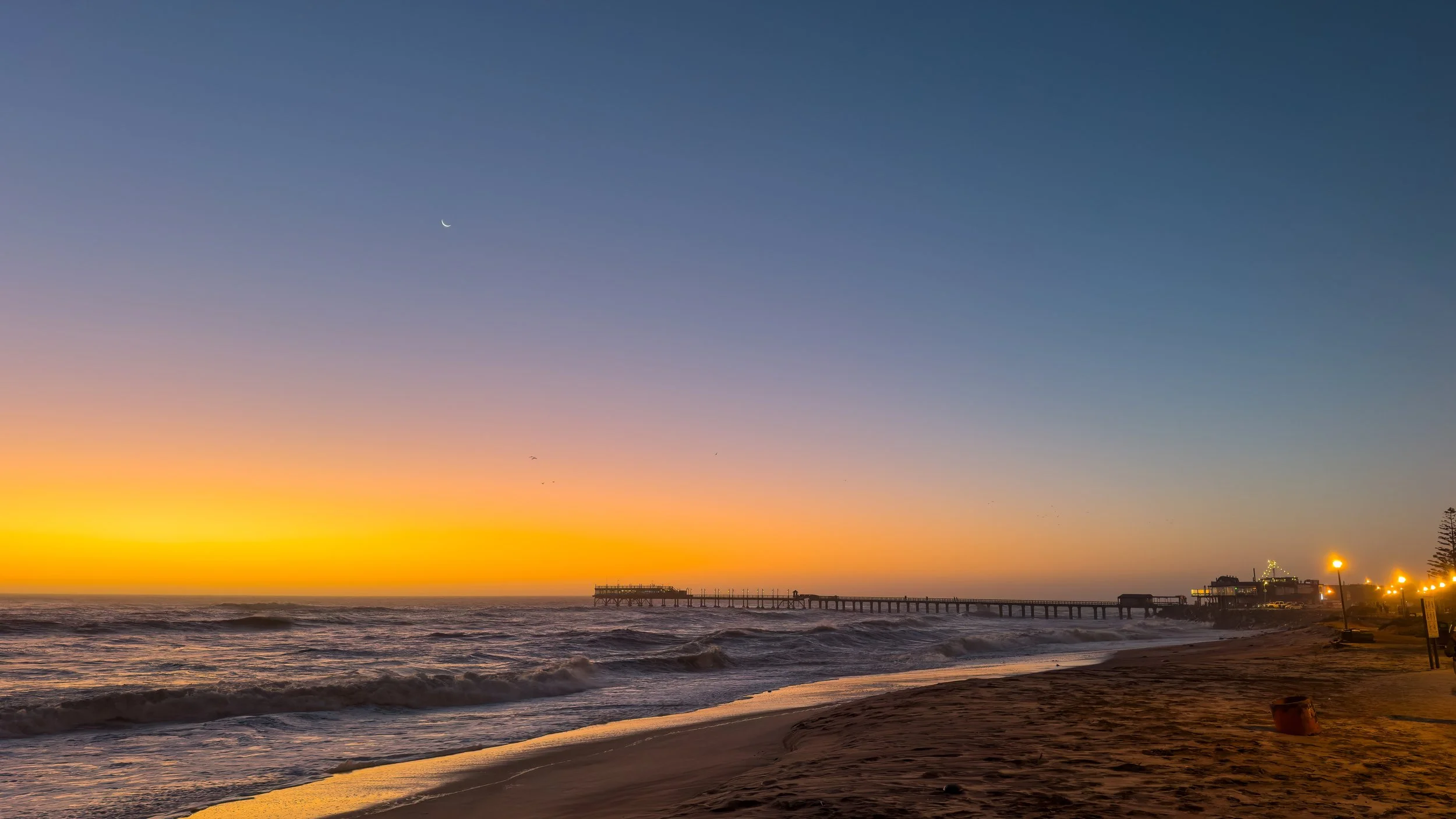 Sunset at the beach with a pier extending into the ocean, colorful sky with blue and orange hues, and a few lights on buildings along the shore.