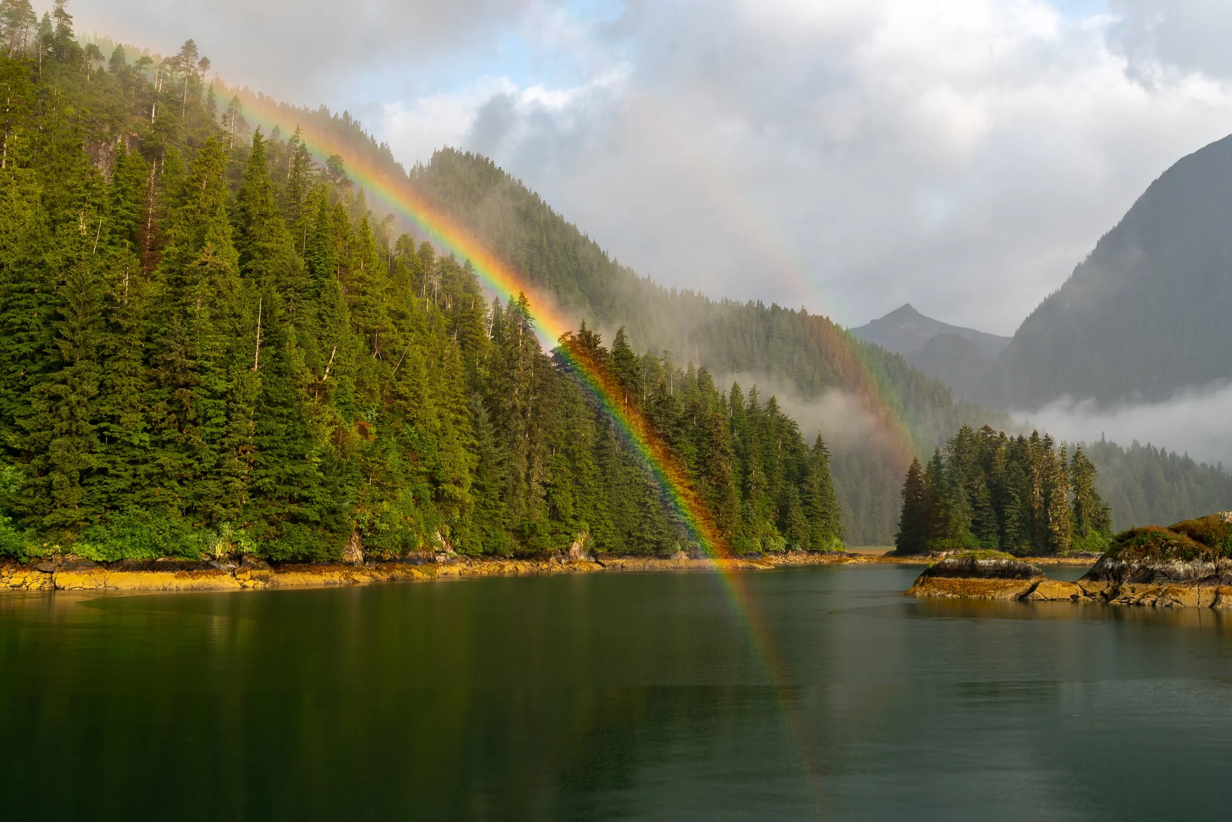 A rainbow arches across the sky, with part of it reflecting in the river at Alaska, USA