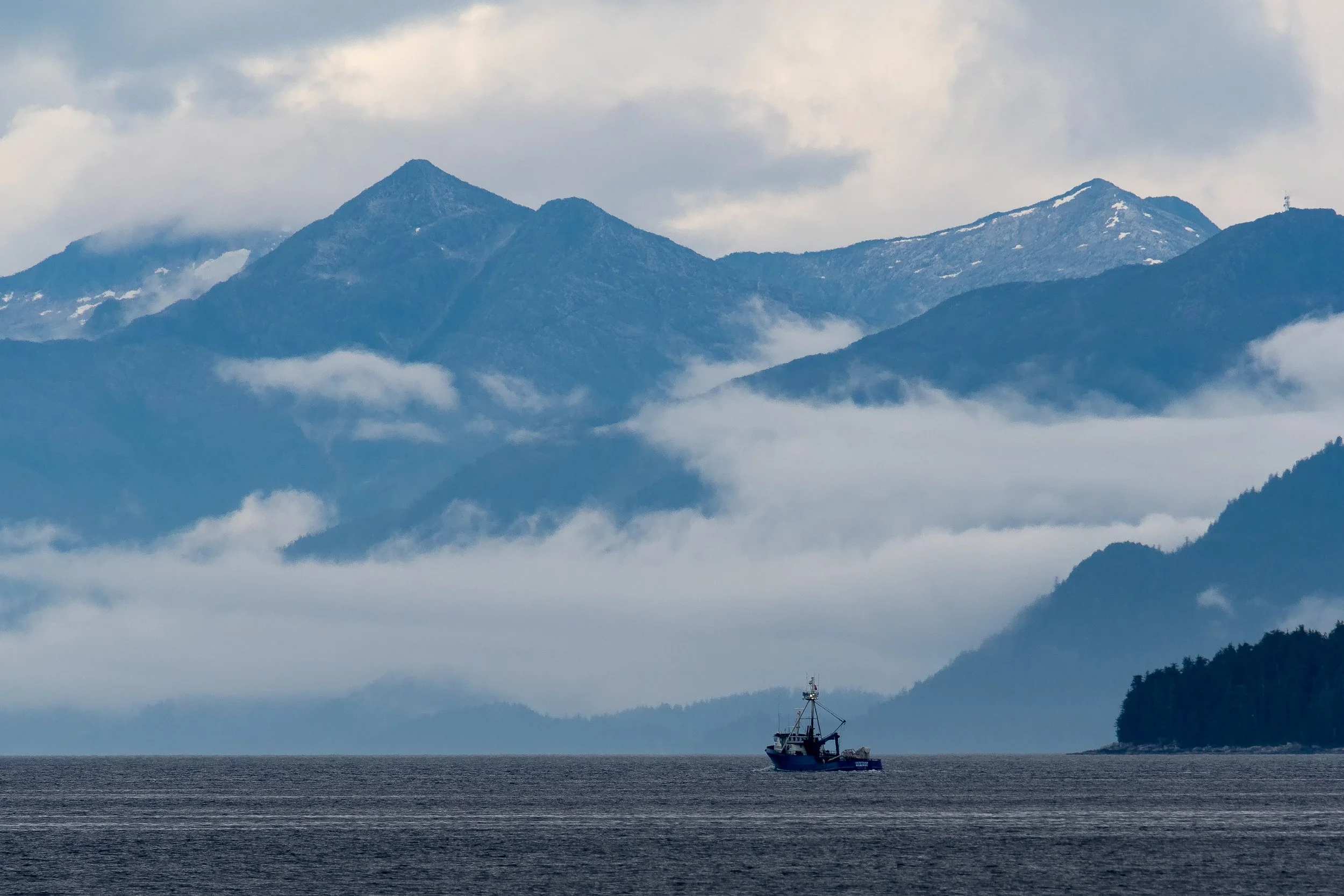 A ship sails a calm bay in Alaska, dwarfed by towering, snow capped mountains , shrouded in low, hanging  clouds.