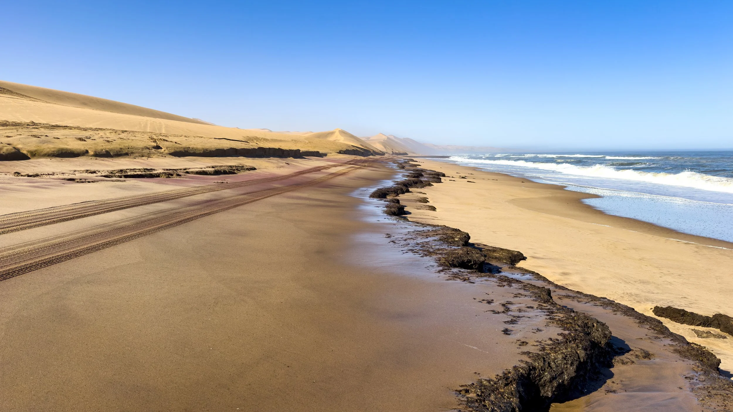 Sandy beach with tire tracks, dark rocks, and ocean waves under a clear blue sky.