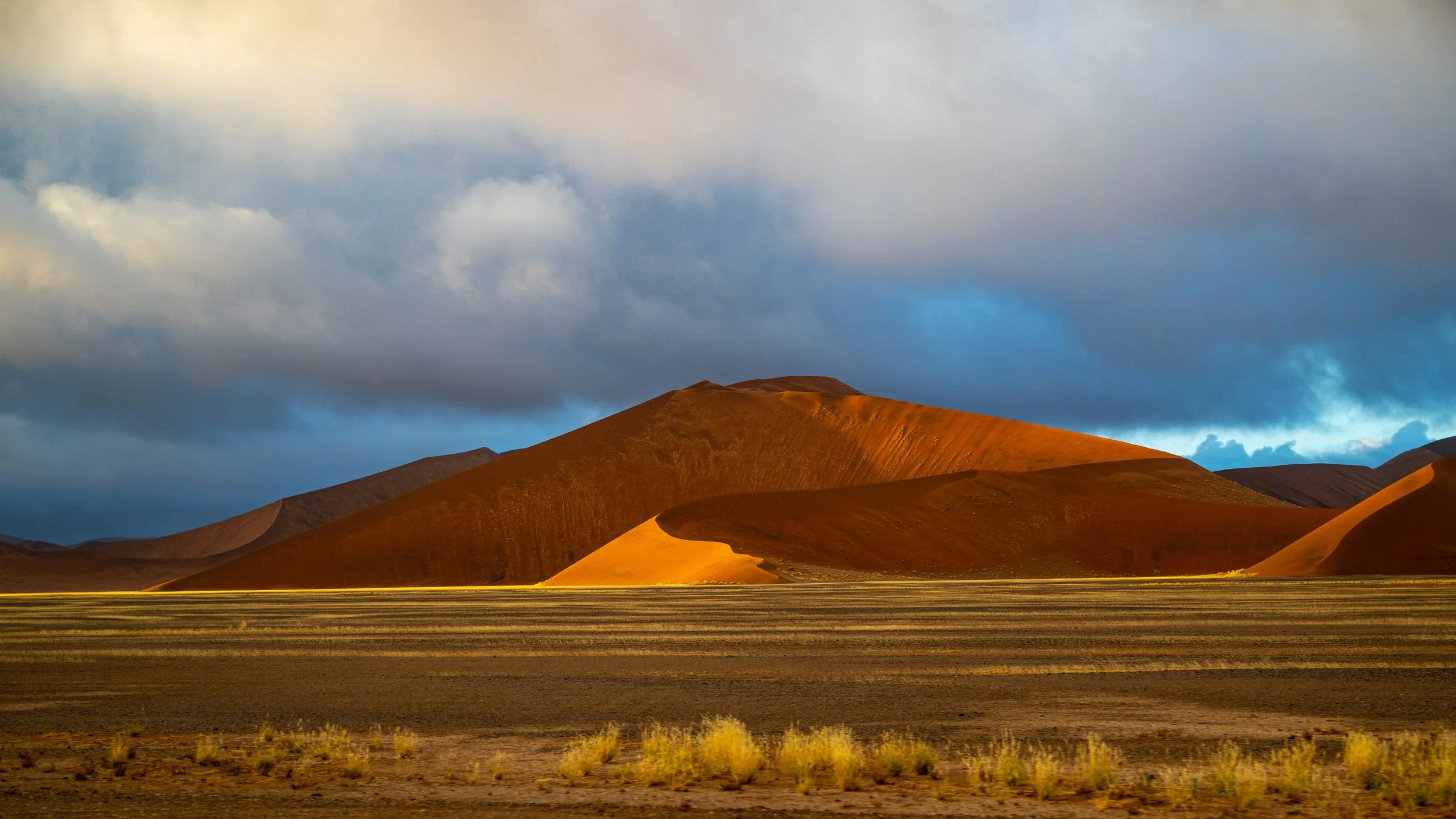 Desert landscape with reddish-brown sand dunes and mountains under a partly cloudy sky