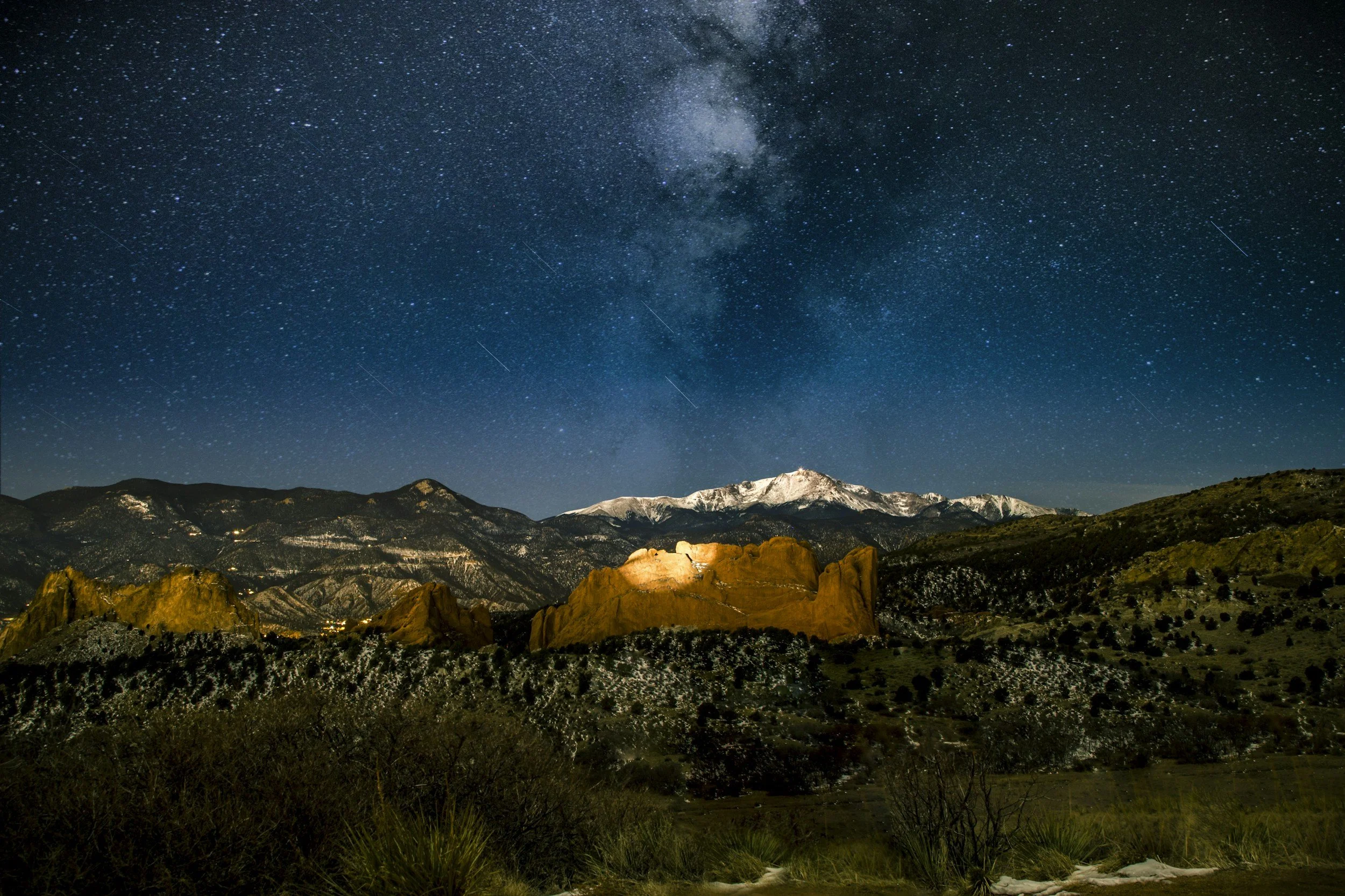 Nighttime landscape with snow-capped mountains, a clear starry sky with the Milky Way, and desert-like formations in the foreground