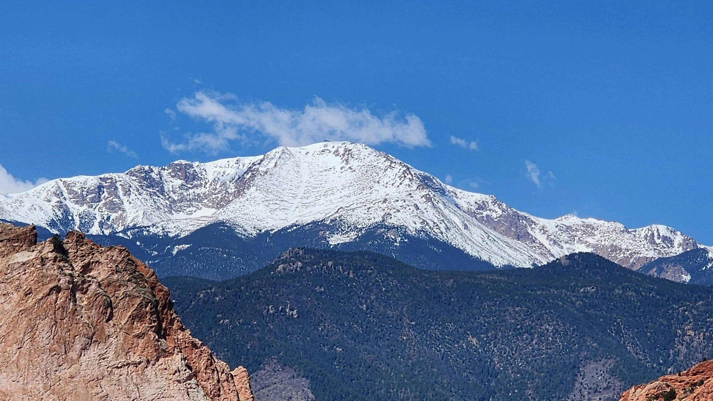 Snow-capped mountain range under a clear blue sky with some clouds, rocky foreground, and forested slopes.