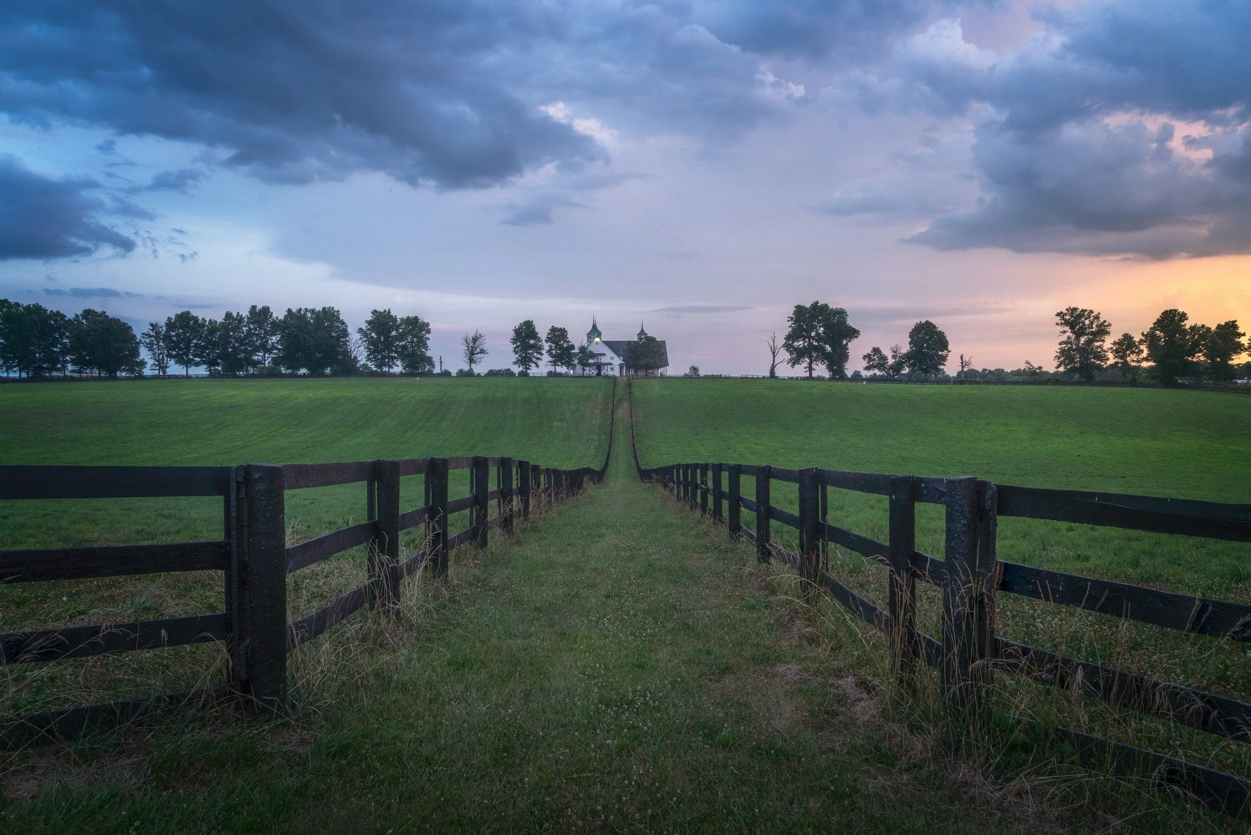 A rural landscape with a grassy field, fenced in with wooden fences, leading to a white church with steeples, surrounded by trees, under a dark, cloudy sky during sunset.