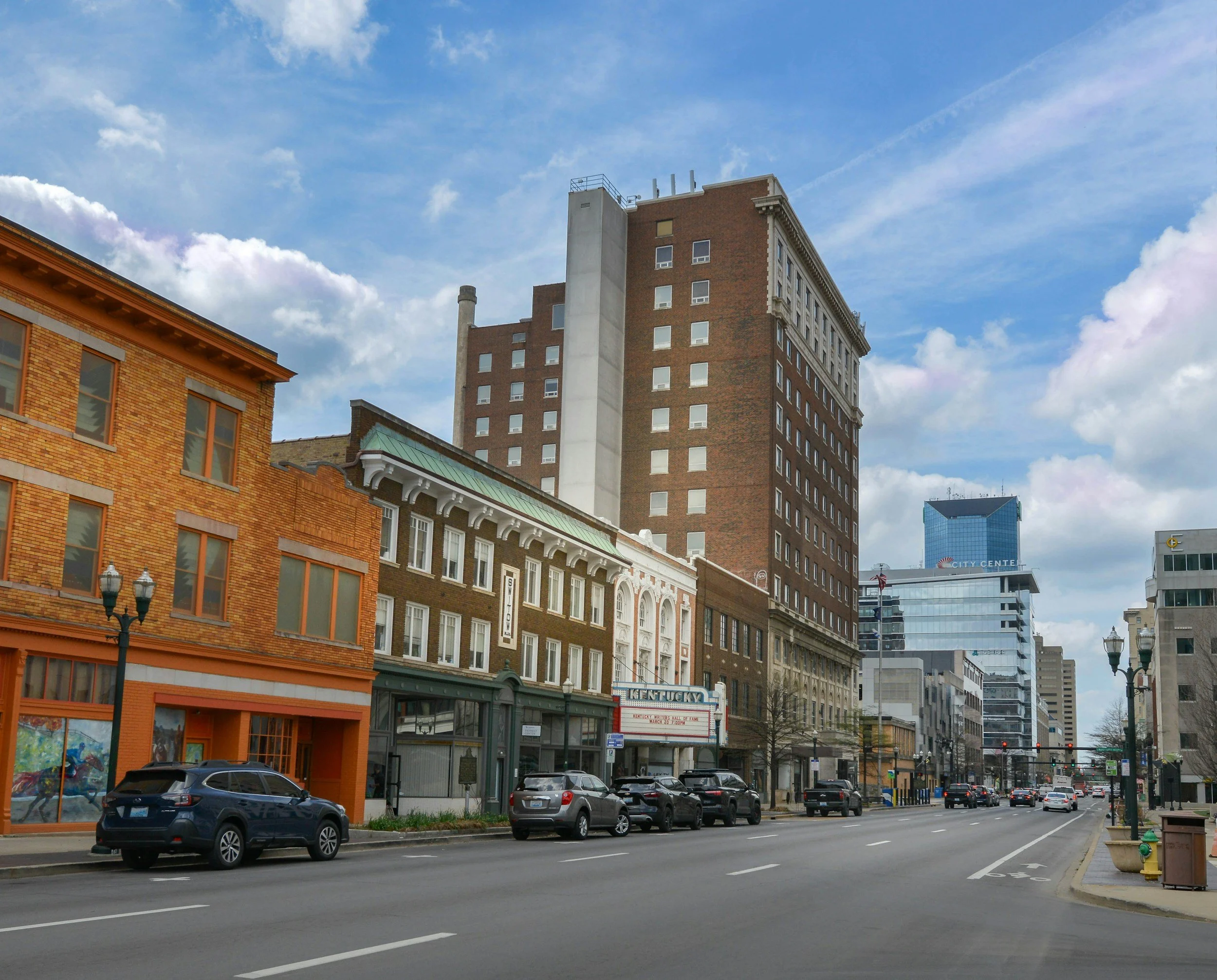 A city street with parked cars along the sidewalk and multistory brick and stone buildings, including a taller high-rise, under a partly cloudy sky.