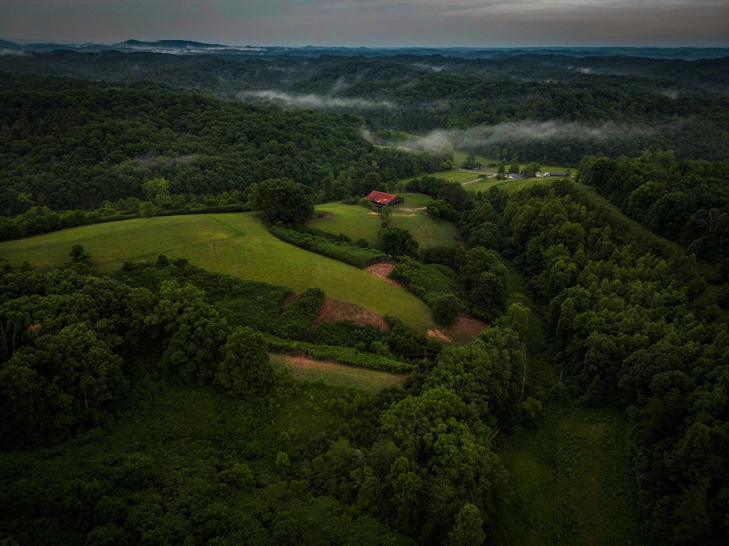 Aerial view of a lush green landscape with rolling hills, dense forests, and a few farm buildings with a red roof, shrouded in mist.