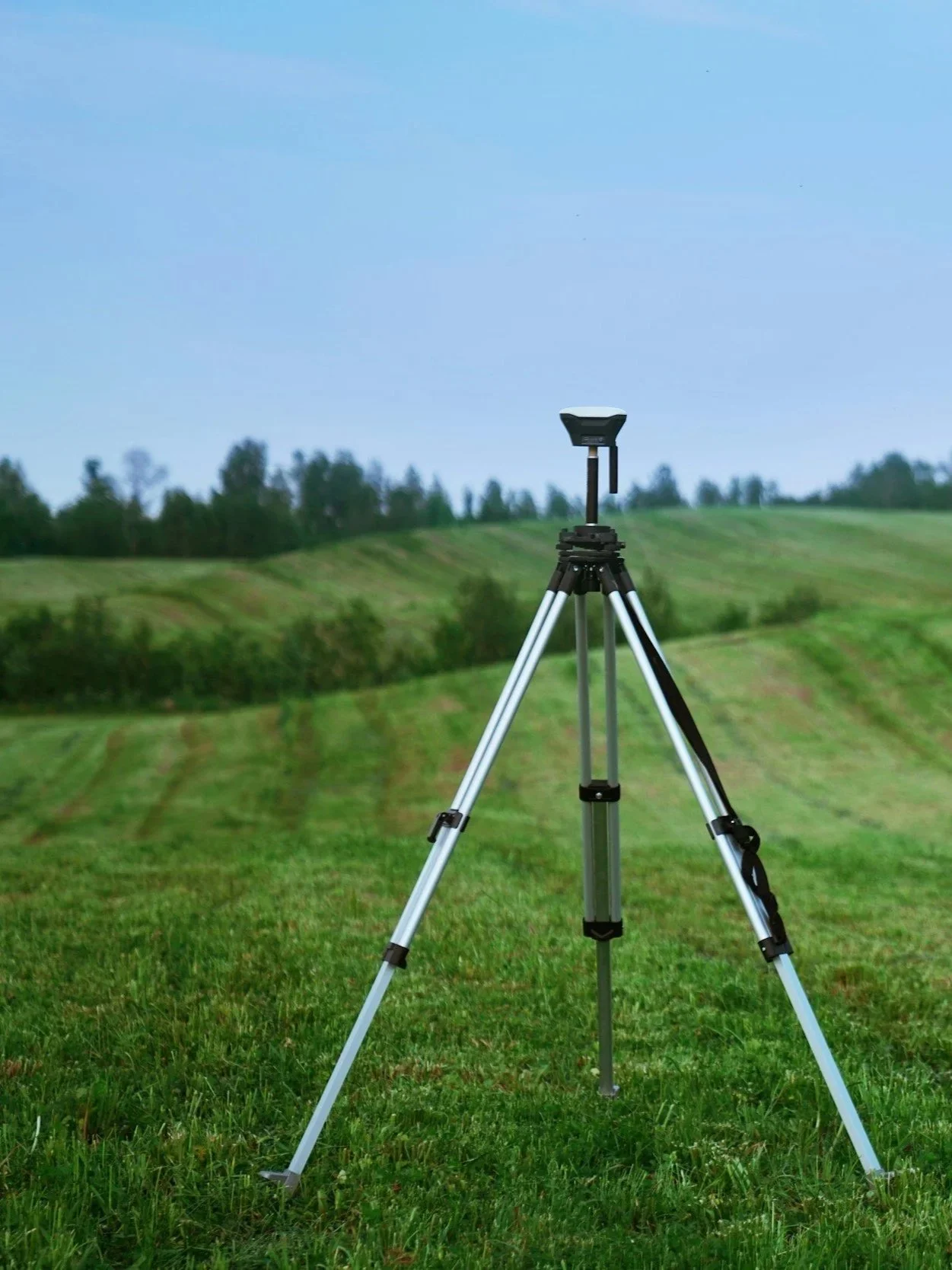 Surveying tripod stand on grassy field with trees and rolling hills in background under blue sky.