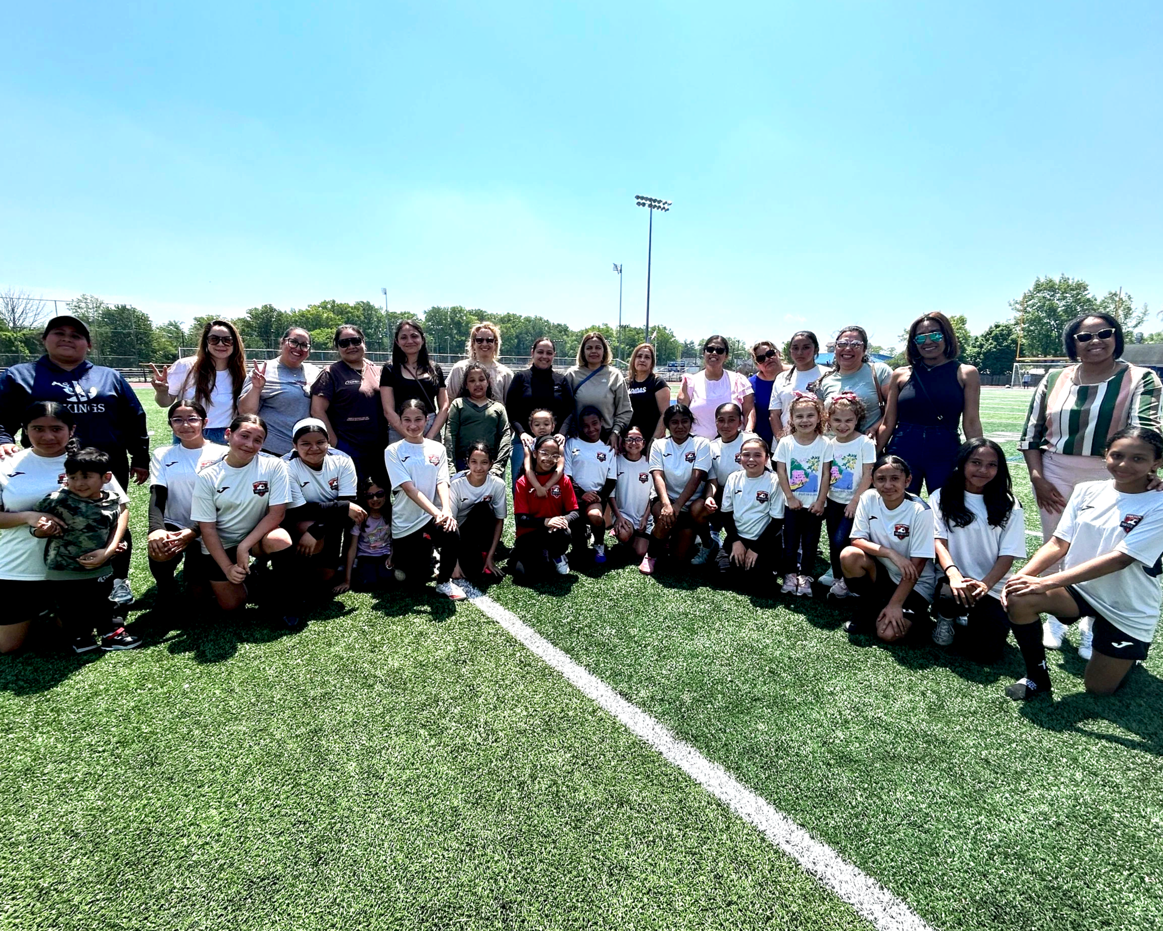 Group photo of children and adults on a football field during daytime, with clear blue sky and trees in the background.