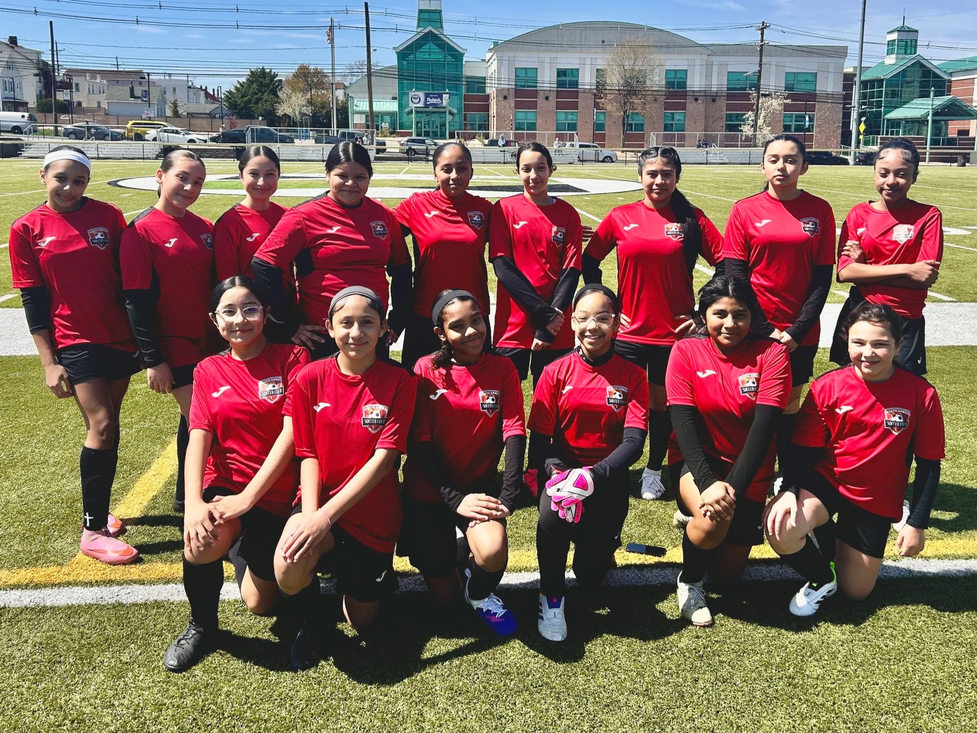 A group of young female soccer players wearing red jerseys and black shorts, posing for a team photo on a soccer field with a school building in the background.