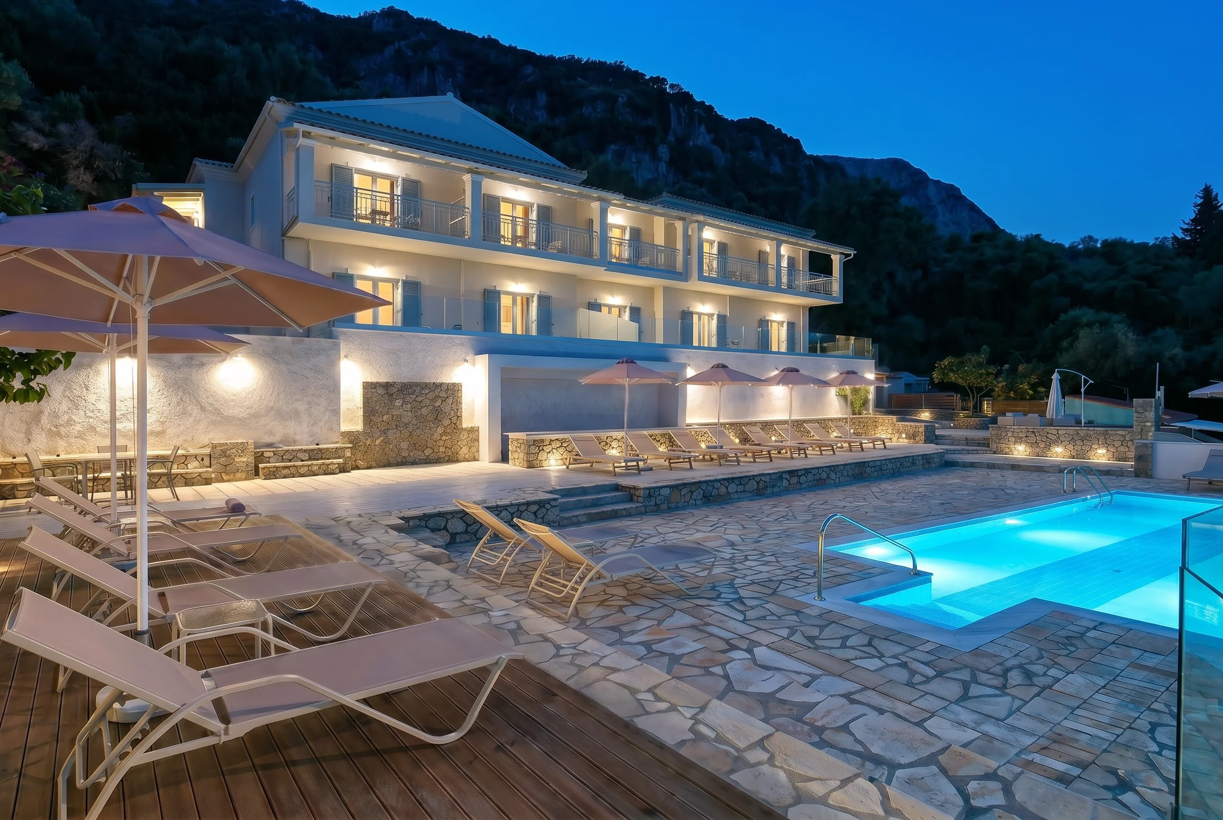 Evening view of Platakia Blu with illuminated pool and oceanfront apartments in Paleokastritsa, Corfu.