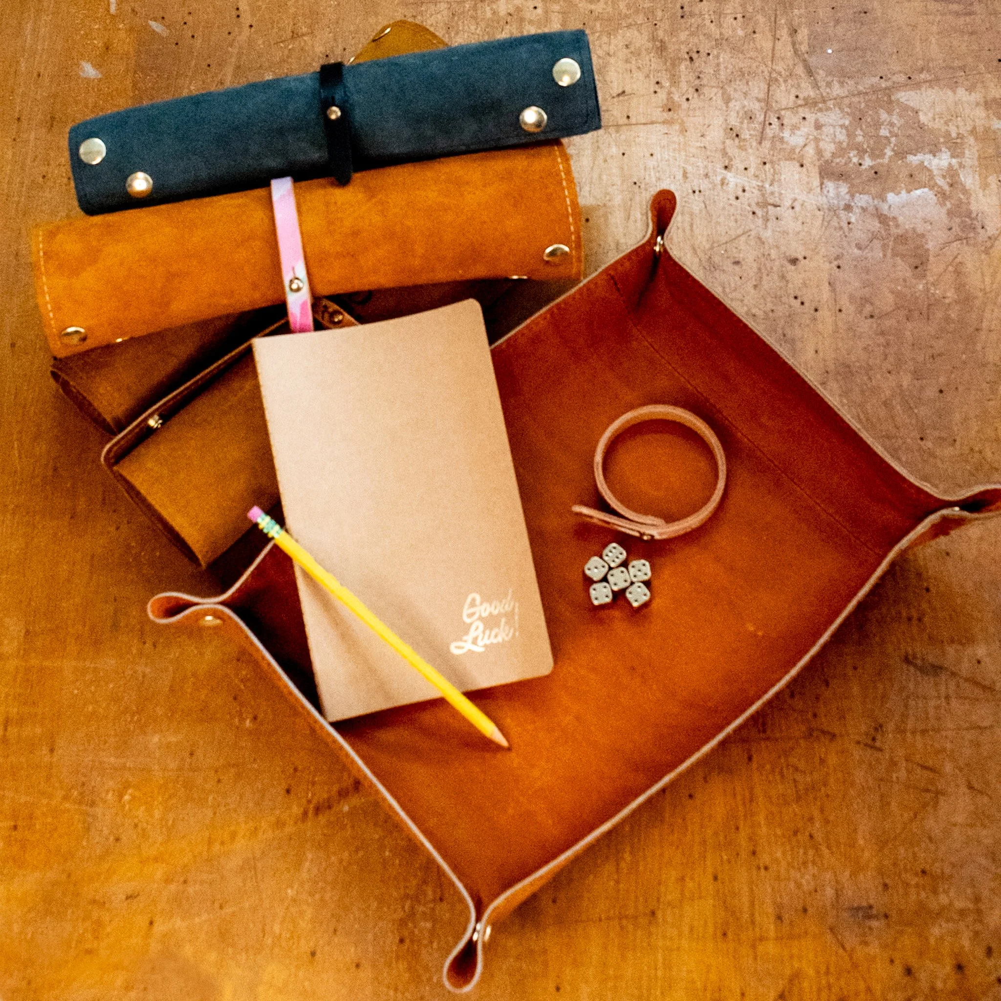 A wooden table holds a brown leather wallet, a black fabric item with metal snaps, a pink and yellow striped pencil, small white dice, and a roll of pink tape.