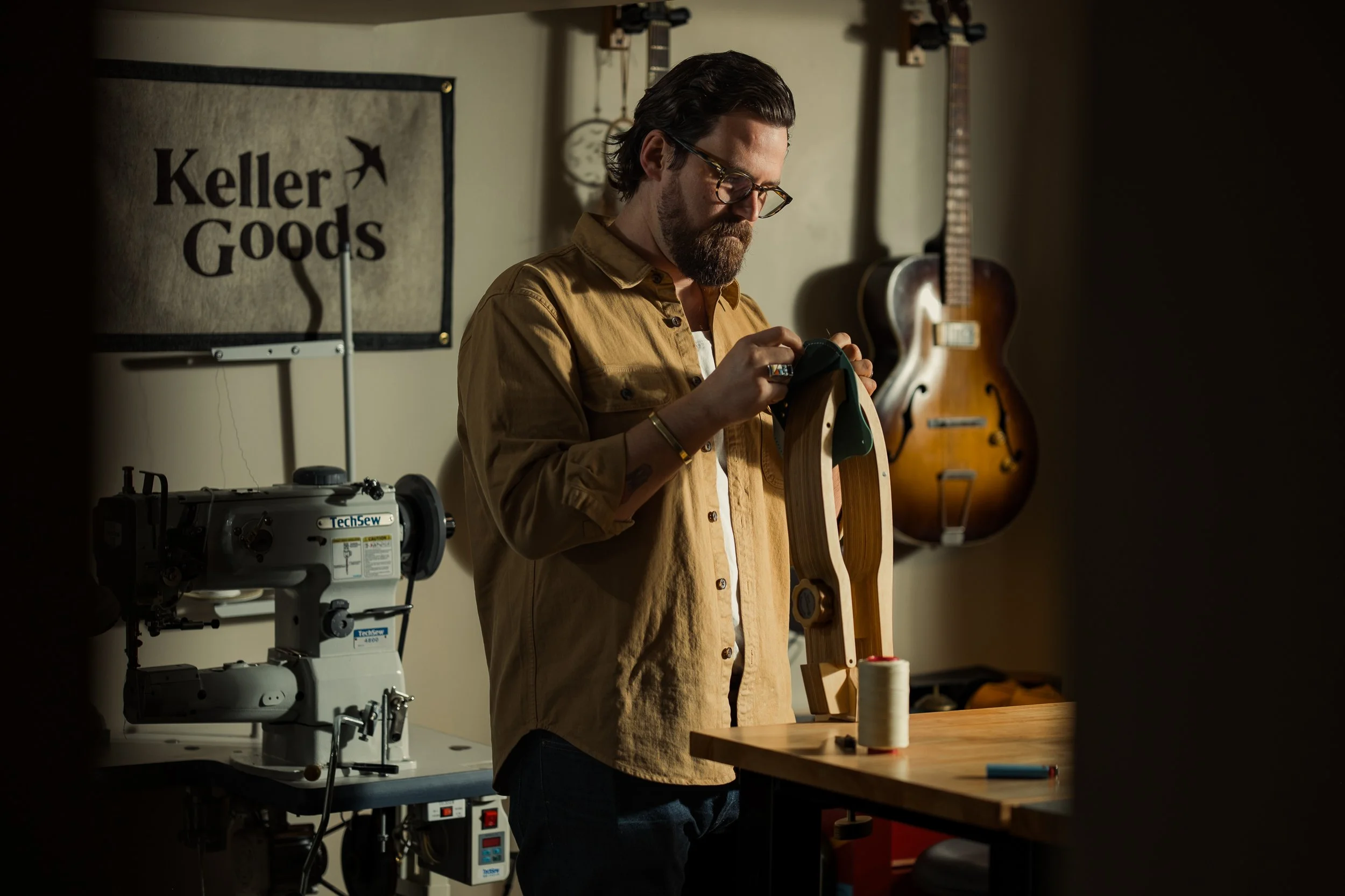A man with glasses and a beard wearing a tan jacket is working on a wooden guitar neck in a workshop. The background features guitars on the wall, a sewing machine, and a sign that reads 'Keller Goods'.