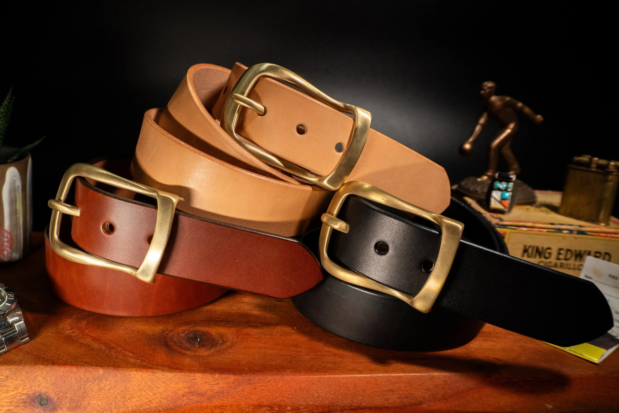 Stack of four leather belts in different colors (tan, black, dark brown, and reddish brown) with gold buckles, resting on a wooden surface with small decorative items and a plant in the background.