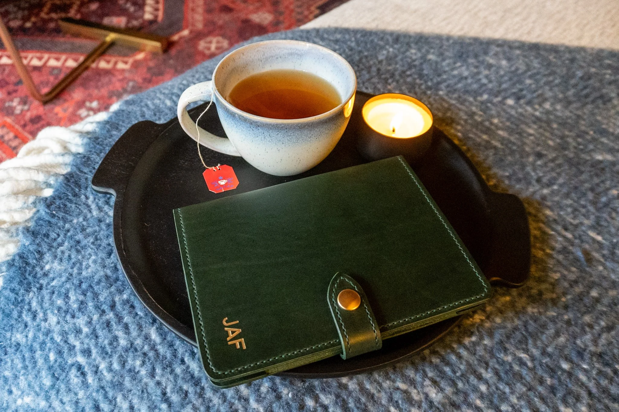A white ceramic mug of tea with a tea bag tag resting on a round black tray alongside a small lit candle in a black holder. The tray is on a dark surface with a green notebook and a leather object nearby, and a red patterned carpet and wooden furnitu