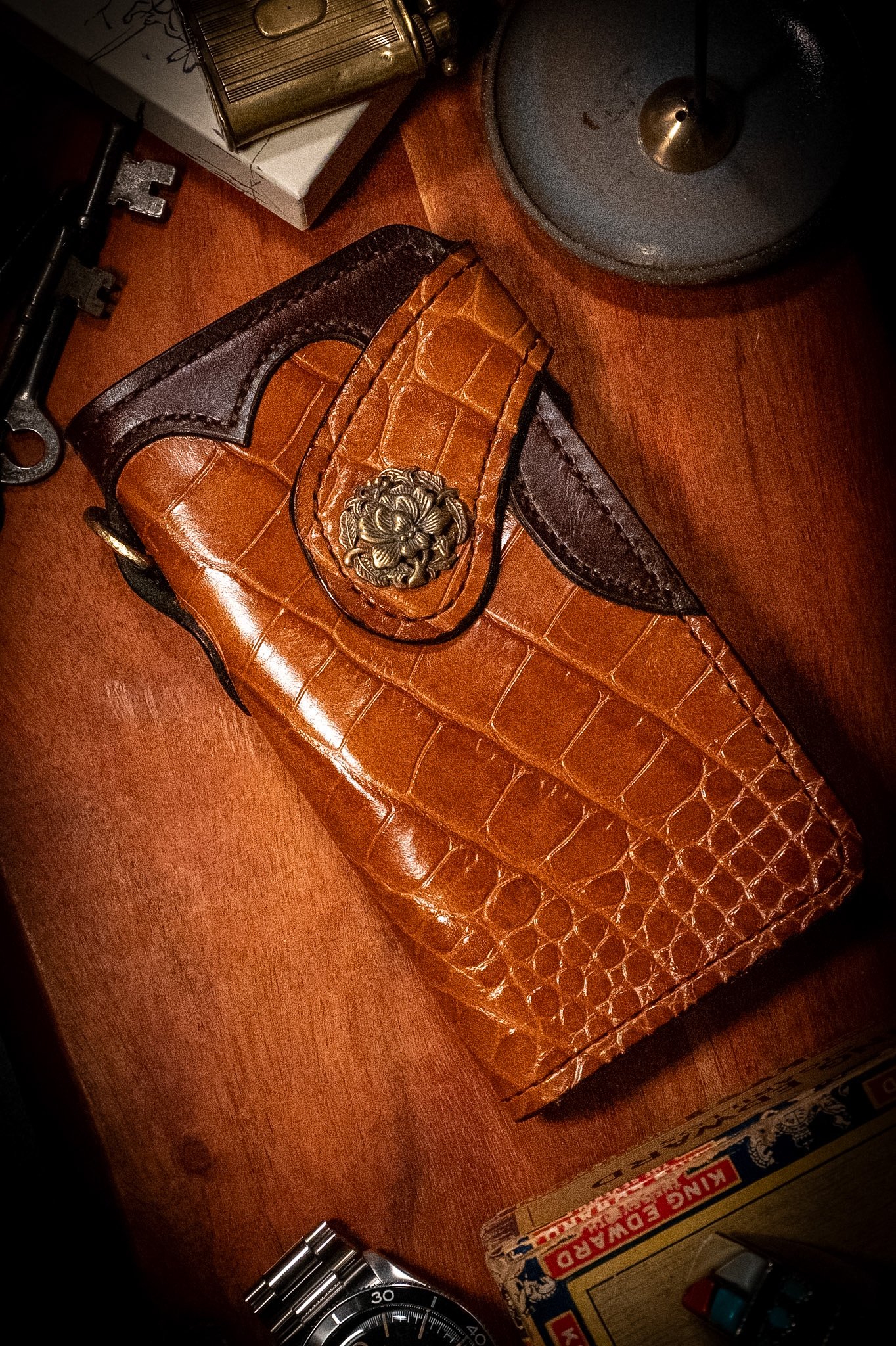 A wooden surface with various objects including a brown crocodile-patterned leather wallet, a metal watch, a metal key, a box of matches, and a box of matches with some small bottles.
