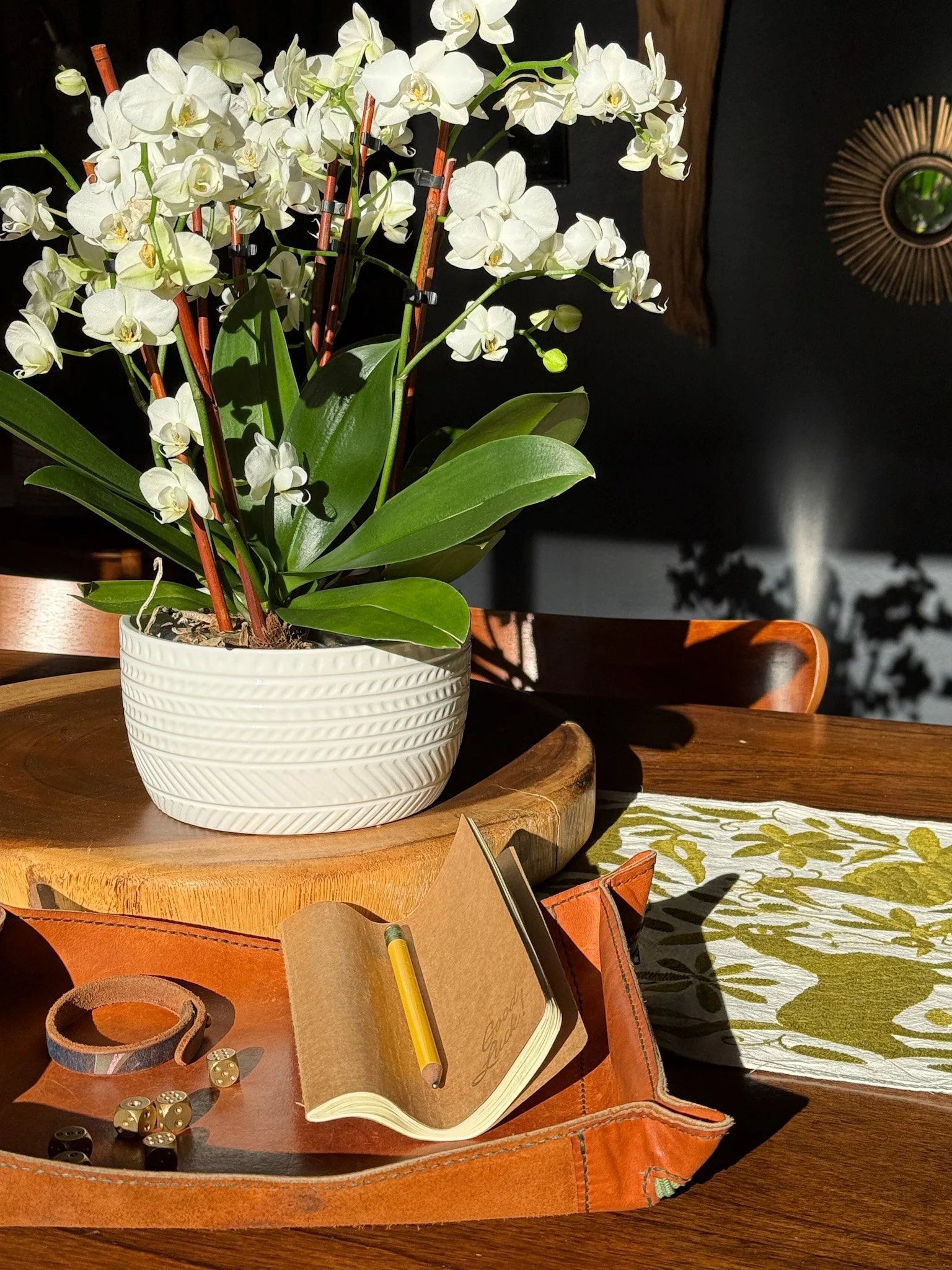 A pot of white orchids on a wooden table with a notebook, dice, and leather bracelet nearby.