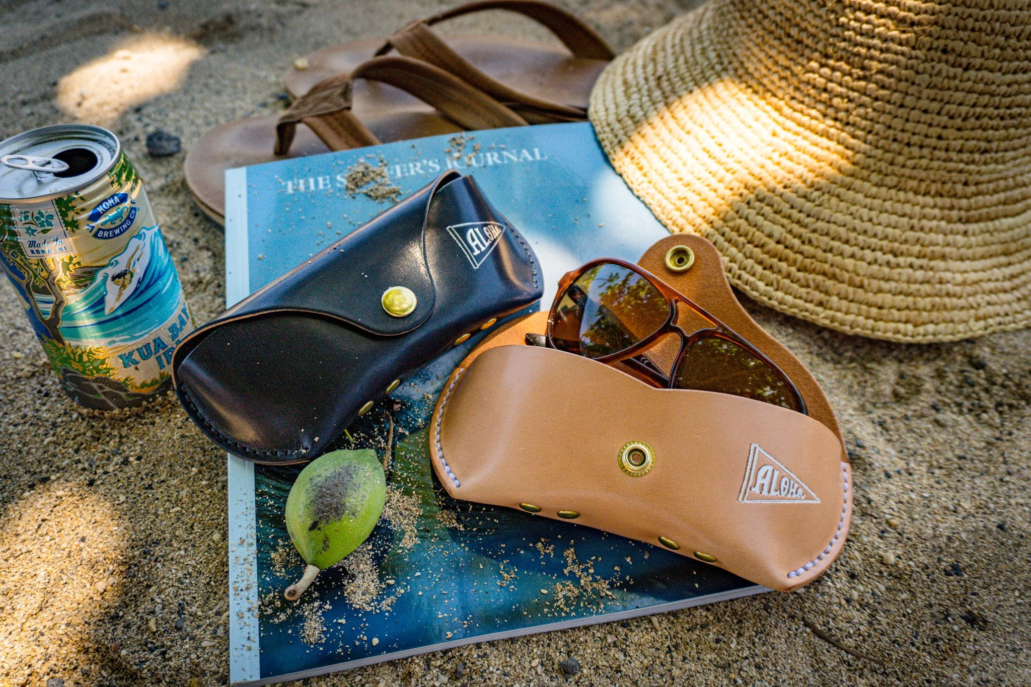 A beach scene with a blue surfboard, a can of Hawaii brewing company soda, a black purse with gold hardware, a pair of sunglasses, a sun hat, and green fruits on sand.