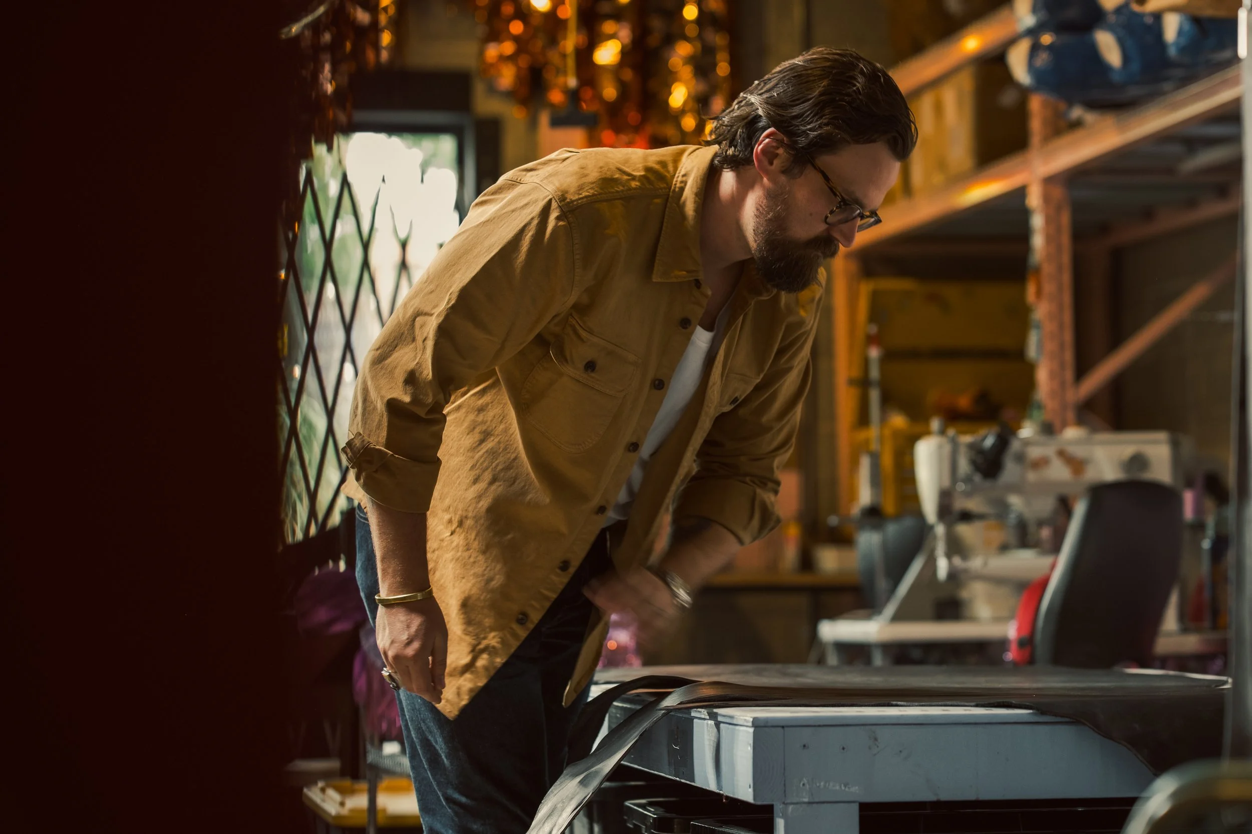 A man with dark hair and glasses is working in a warehouse or workshop, wearing a tan jacket and leaning over a table with equipment or tools.