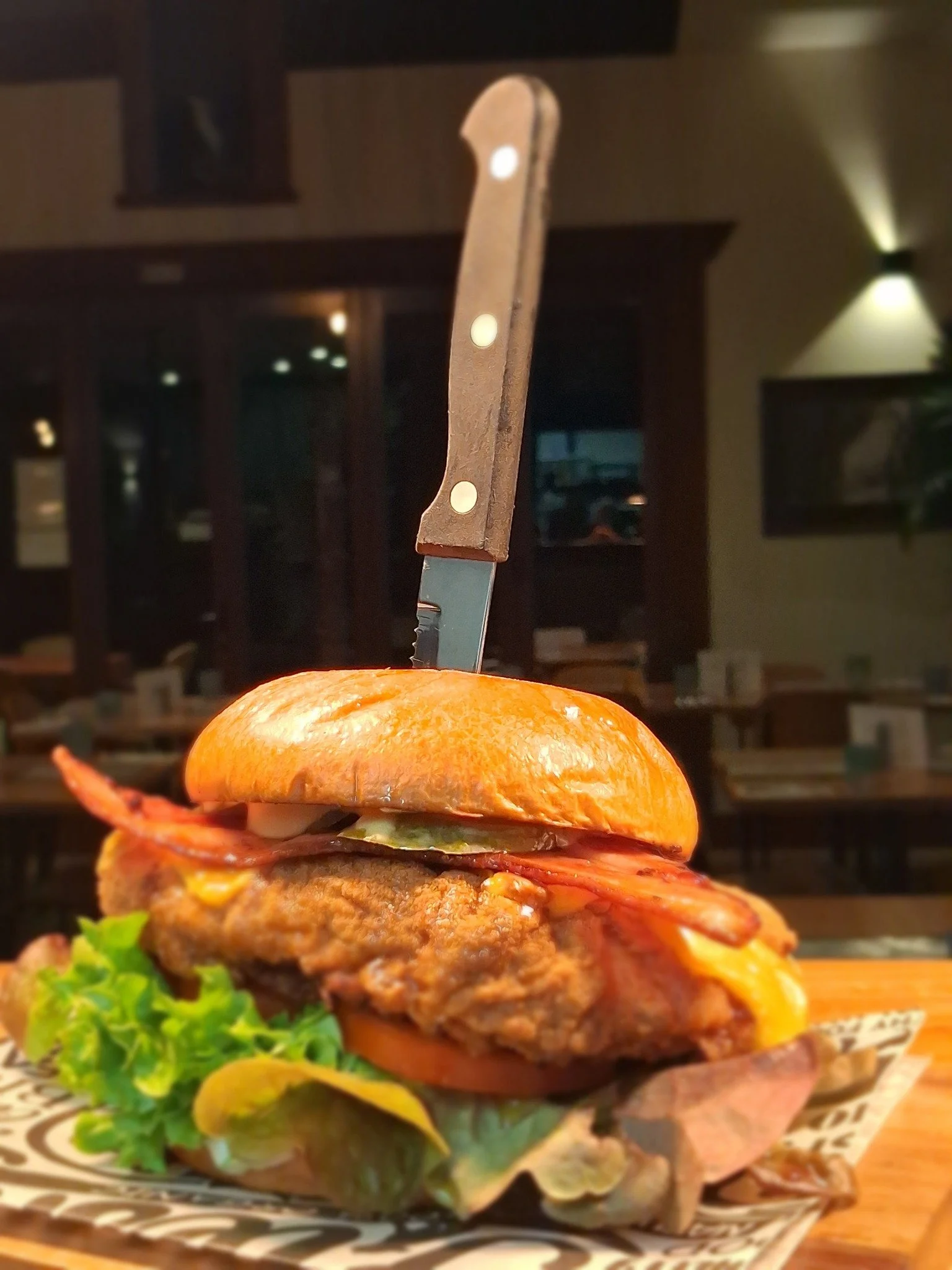 A close-up of a gourmet burger with a toasted bun, lettuce, tomato, a beef patty, cheese, bacon, and a slice of pickles. The burger is topped with a knife inserted into the bun, set on a decorative plate with a leafy garnish in the foreground.
