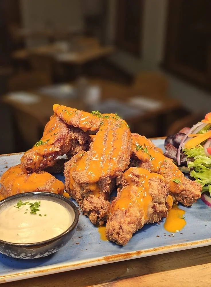 Fried chicken wings drizzled with orange sauce, served with a side of creamy dipping sauce and a small salad, on a rectangular blue plate in a restaurant setting.